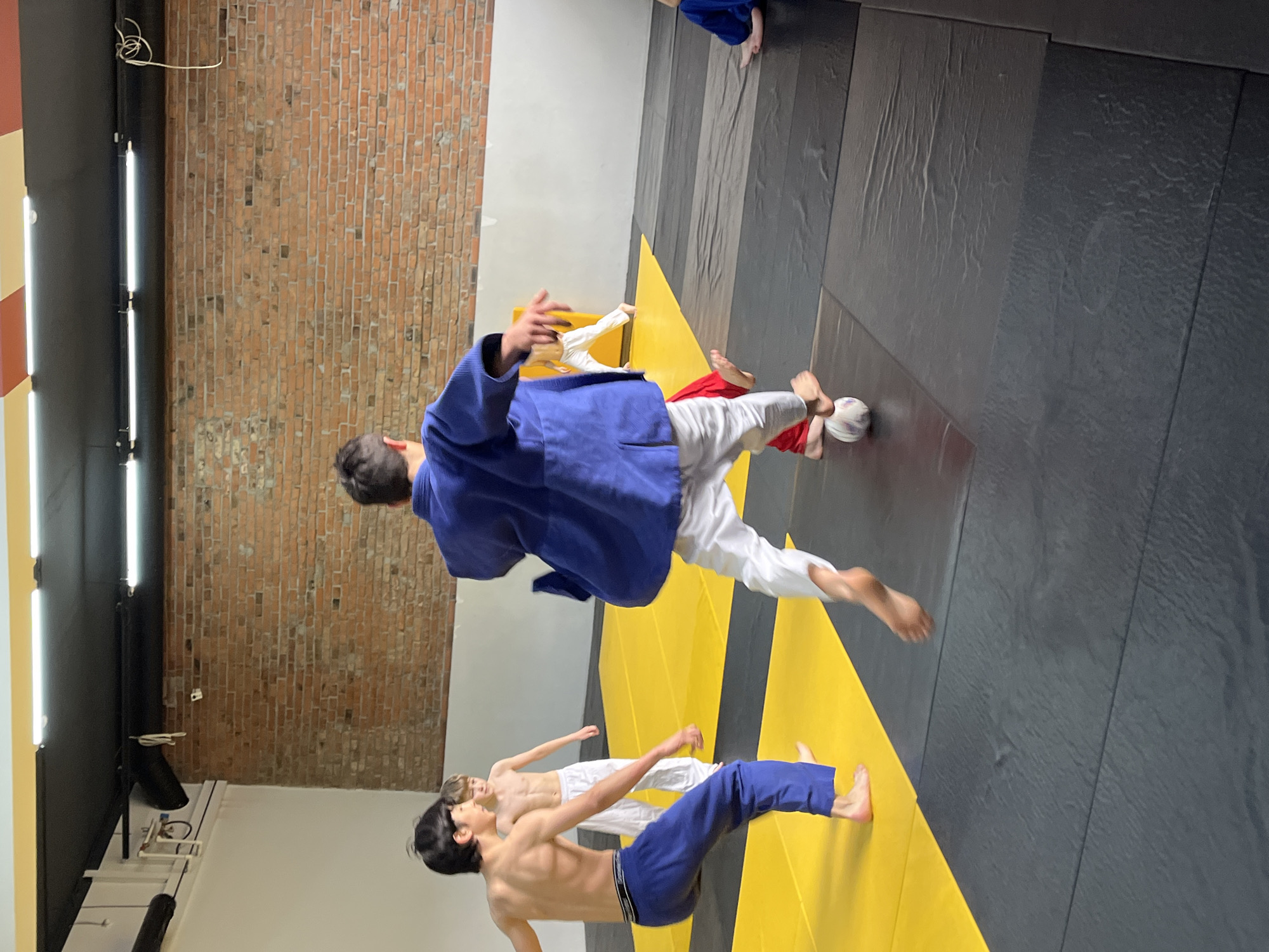 The image depicts a judo training session in what appears to be a gymnasium or martial arts training facility. Several individuals, likely judokas or practitioners of a similar combat sport, are seen practicing techniques on a matted surface. The mats consist of alternating yellow and dark gray panels. One individual is wearing a blue judogi (uniform) and white pants, appearing to be in mid-air, potentially executing a throw or takedown. Another individual, bare-chested, is wearing white pants and blue uniform bottoms. In the background, a brick wall is visible, along with lighting fixtures. Based on the available information, it is not possible to confirm with certainty the location in the city of Krasnoyarsk, Russia.