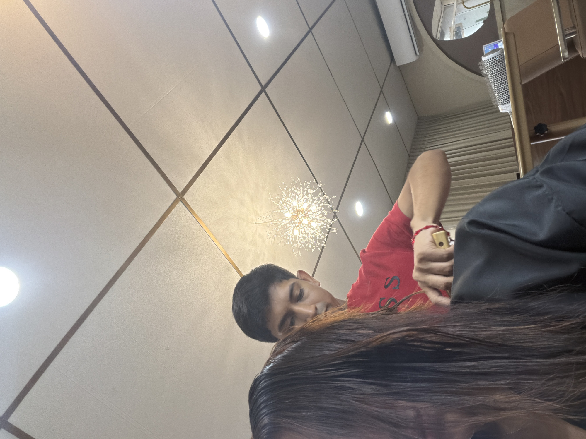 A medium shot depicts a man with dark hair, wearing a red t-shirt and a red bracelet, cutting long brown hair. The setting appears to be indoors, possibly a hair salon, under bright artificial lighting. The walls are beige with thin brown lines forming squares, and a decorative light fixture is visible. The scene suggests a hair styling session in progress. Location is in Villahermosa, Mexico.