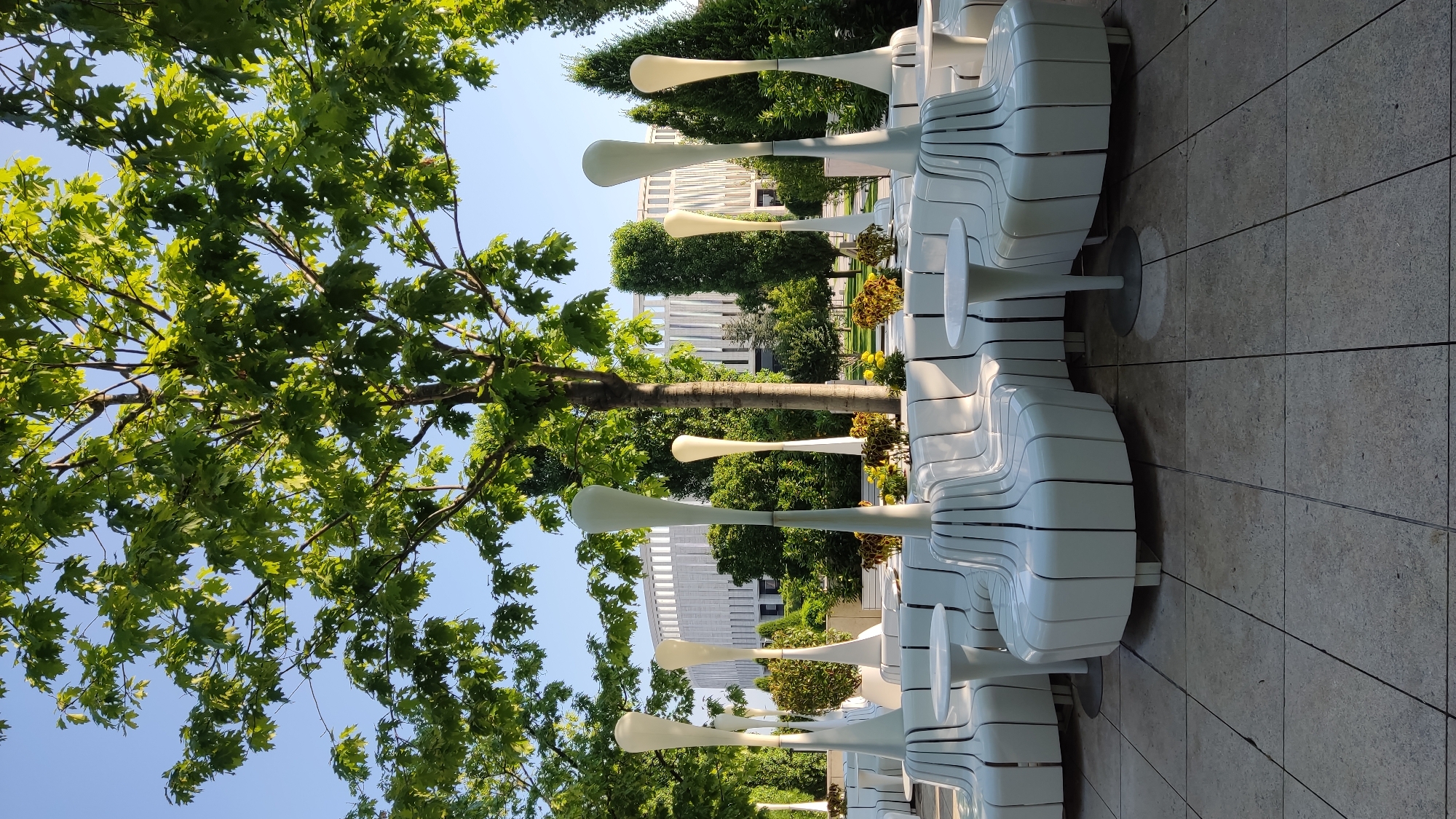 The image depicts an outdoor seating area with a modern design, likely located in Krasnodar, Russia. In the foreground, there are curved, white benches with a slatted design, arranged in a semi-circular formation around small, white, pedestal tables. Tall, white, decorative lamps with a bulbous top stand intermittently behind the benches. Behind the seating area, there is lush greenery with various trees and shrubs, and in the background, a large, modern building with a distinctive facade is visible. The sky is clear and blue. The scene suggests a public park or a commercial establishment with an emphasis on contemporary aesthetics. No people are present in the image. The ground is paved with square tiles.