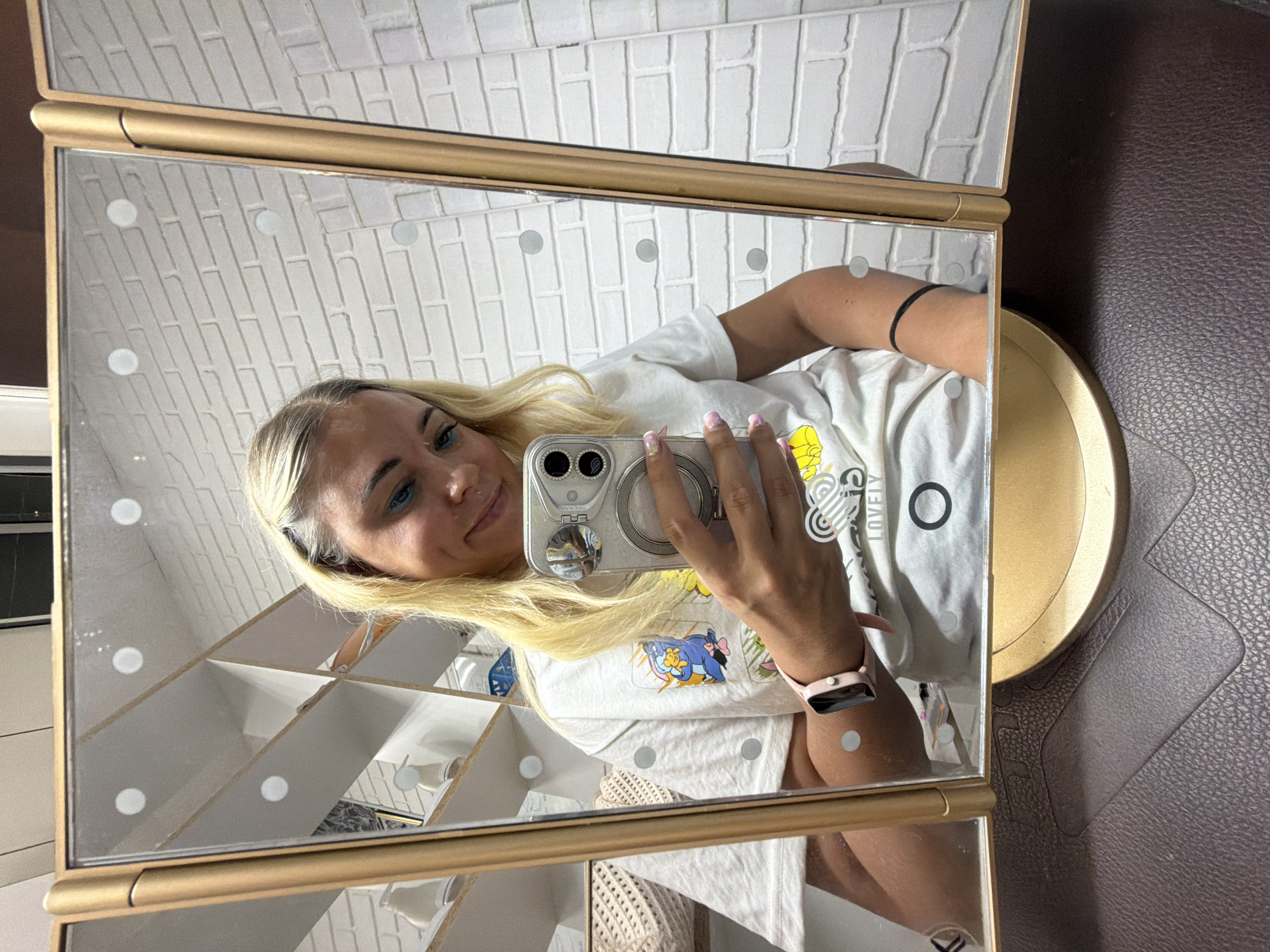 The image shows a young woman with blonde hair taking a selfie in a tri-fold mirror. She is wearing a white t-shirt with cartoon characters and text on it. She has blue eyeshadow and light pink nail polish. Her left arm, visible in the mirror, has a black band. She is also wearing a pink smartwatch. The mirror is framed in gold and has integrated lights. Reflected in the mirror is a white brick wall and shelves. The background is a dark brown, textured surface, possibly leather. The overall impression is casual and personal. The image is oriented vertically.