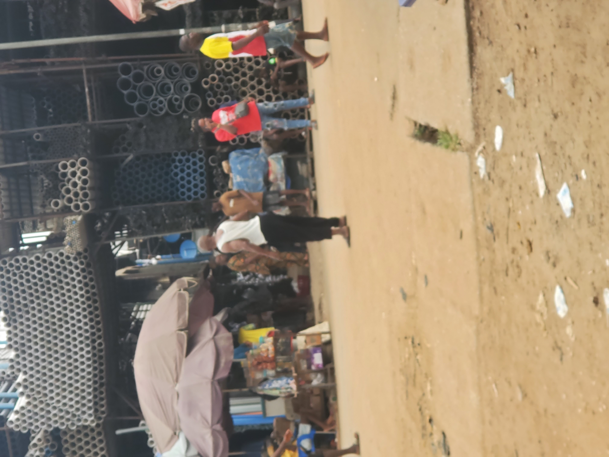 In Fegge, Nigeria, a street scene displays commerce and daily life. Stacks of PVC pipes form the backdrop on one side, indicating a hardware business. People casually walk along the dusty ground; they are of different sizes and dress casually. An umbrella shades a small market stall displaying assorted goods. The ground shows signs of foot traffic and scattered debris.