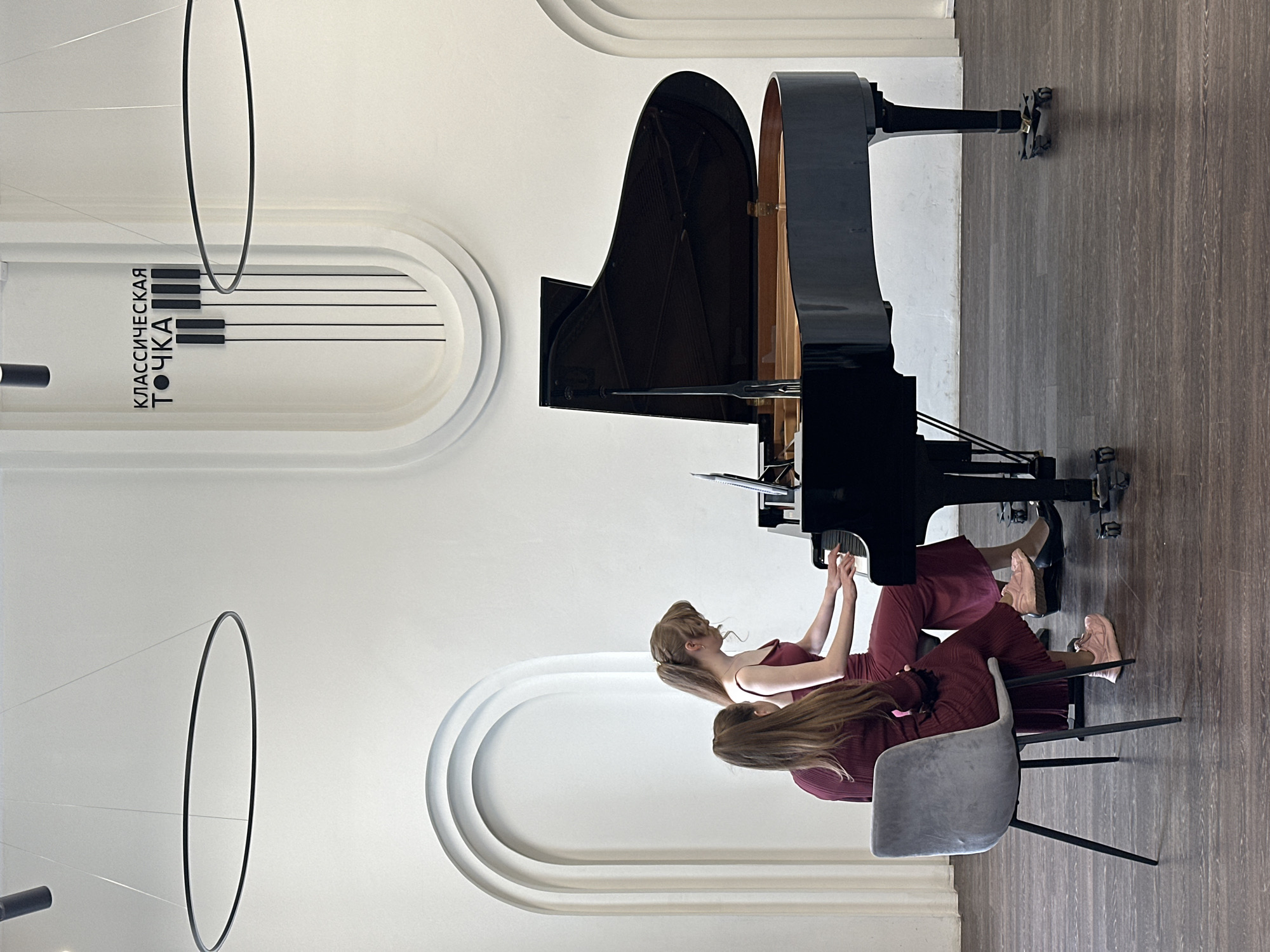 Two women sit at a grand piano. The piano is black and has its lid open. The women are wearing maroon dresses and the woman in front is wearing pink sneakers. The background includes a white wall with a sign in Russian that translates to "Classical Point." Above the piano are two circular light fixtures. The floor is dark wood. The location appears to be in a concert hall or music venue.