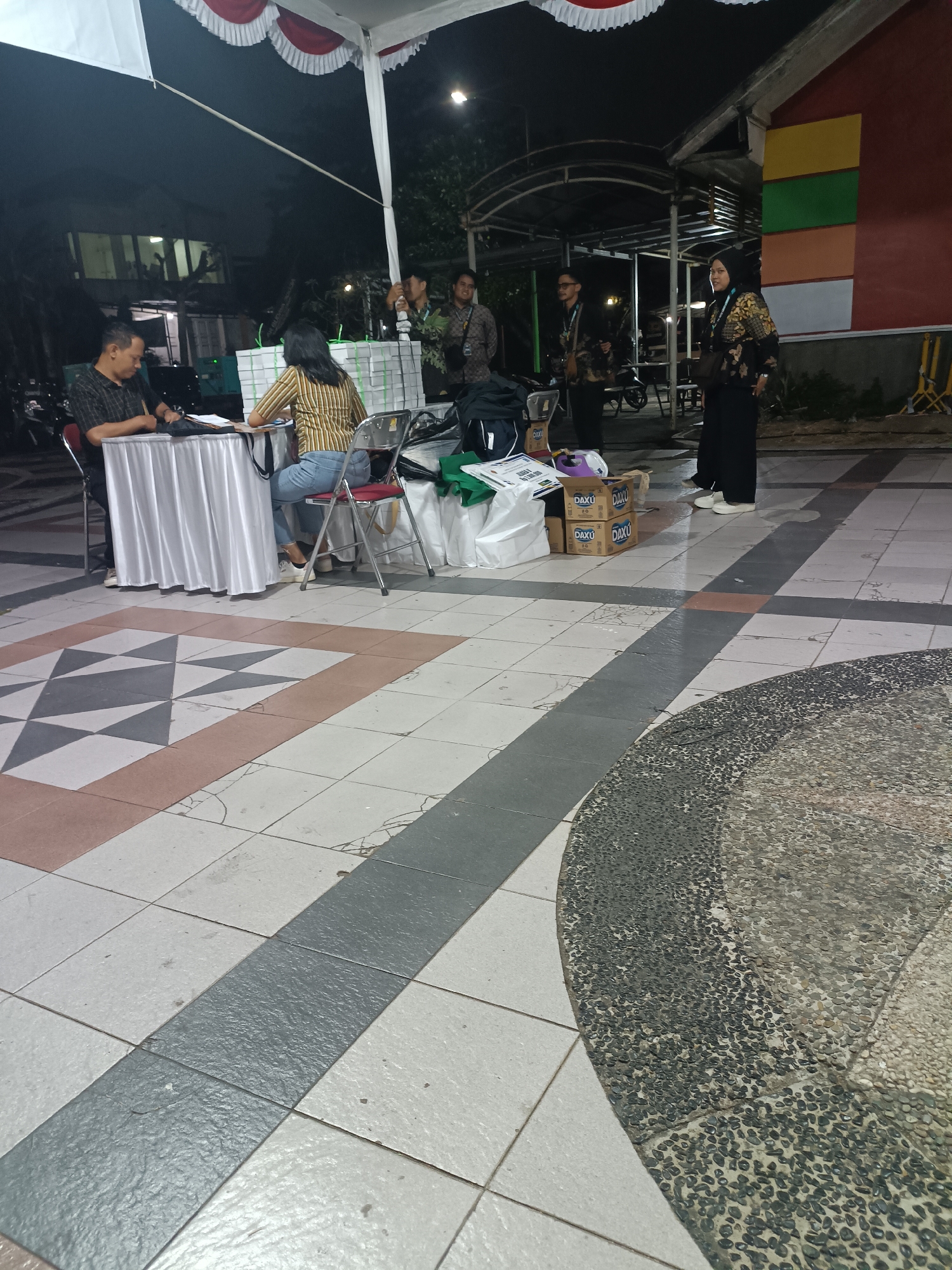 The image depicts an outdoor event space in Condongcatur, Indonesia, likely at night given the lighting. In the foreground, a tiled walkway with a geometric pattern is visible. Several people are present, some interacting at a table covered with a white cloth. One man is seated, looking down at papers, while a woman in a striped shirt is seated opposite him. Behind them, a stack of white boxes with green writing is present. Further back, several other individuals, some in dark clothing with patterned accents, are standing. The area is partially covered by a white tent with red and white trim. The background shows a building with colorful rectangular panels and some trees and vegetation. The overall scene suggests an administrative or registration area for an event.