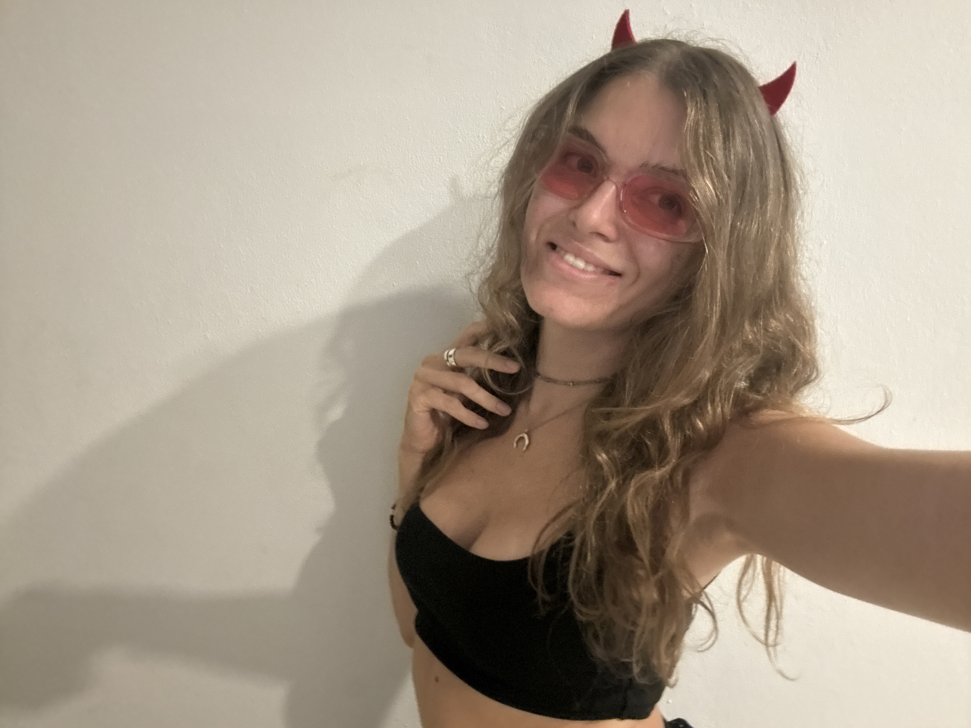 A young woman stands indoors, likely against a plain white wall, in a well-lit room. She is wearing red devil horns, red sunglasses, and a black tube top. She has shoulder-length brown hair and is smiling. She wears jewelry, including a ring and a necklace. Her right hand is raised towards her face, and her left arm is extended, suggesting she is taking a selfie. There's a shadow of her face and hand on the wall behind her. The location, Moo 4, Thailand, is not discernible from the image itself.