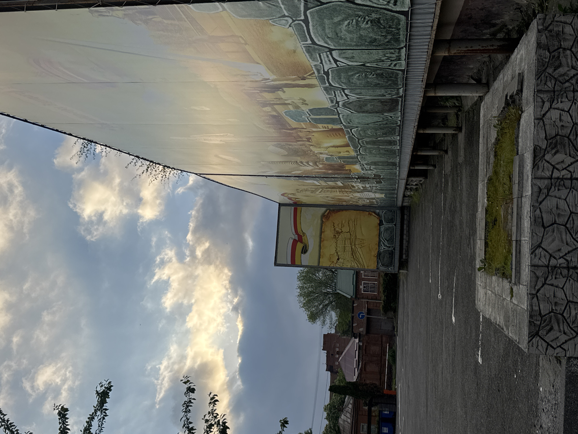 The image shows a parking lot in Akhieli, Georgia, adjacent to a large billboard and a stone wall. The billboard is elevated, with a design resembling a historical panorama and a smaller sign beneath depicting a map or historical document. Buildings, including a brick structure, are visible behind the billboard. The sky is partially cloudy. The parking lot is marked with white lines, and a section of the stone wall is topped with green vegetation.
