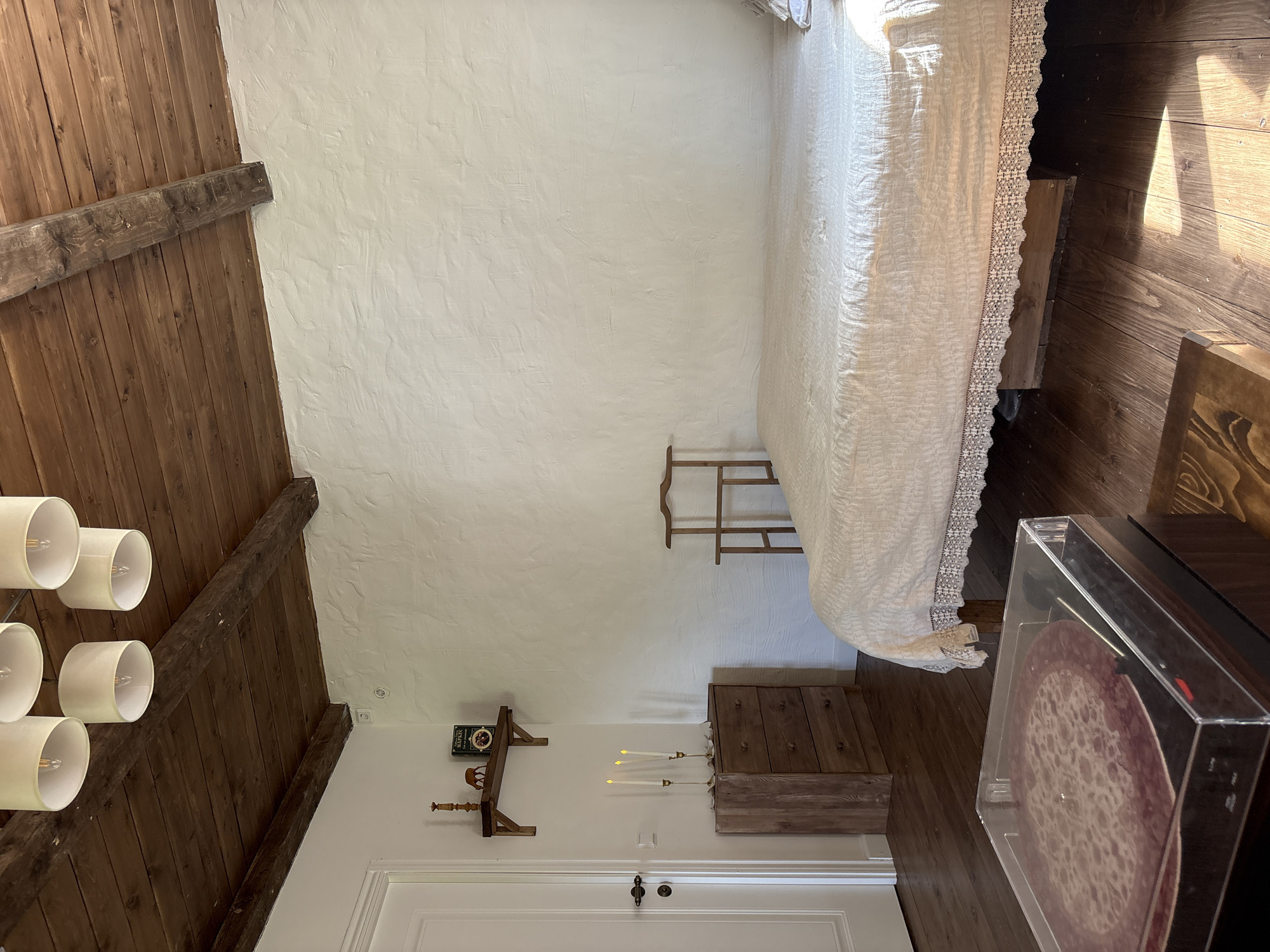 A tilted, indoor shot presents a bedroom with distinct features. The roof and a portion of a wall are made of wooden planks, contrasted by a white stucco-like finish on another wall. A bed with a textured white bedspread occupies the upper right of the frame. Beneath the bed's edge, a small, wooden stool is visible.
On the lower part of the wall, there is a shelf with objects including a book and a decorative item. Further down, a set of faux candles sits atop a wooden chest of drawers. Below this, a white door with a black handle is seen.
A portion of a modern record player is in the bottom right corner, obscuring part of the floor, which appears to be made of wooden planks. Suspended from the ceiling in the upper left are several modern light fixtures with cylindrical, white shades. The setting appears to be a bedroom. There is no clear indication of the location within Sibir', Russia.