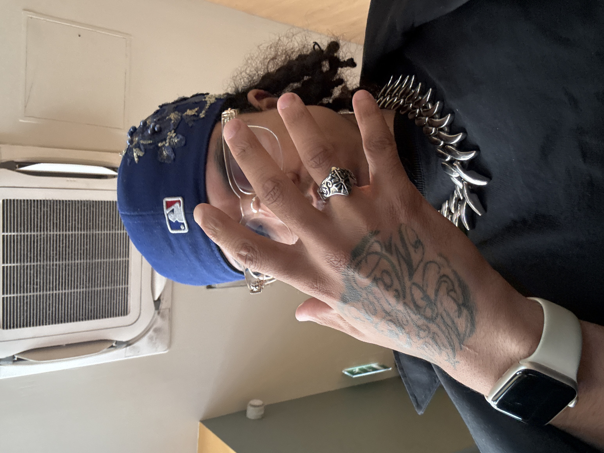 A person wearing a blue baseball cap with floral decoration, eyeglasses, rings, a silver necklace, a black shirt, and an Apple Watch is shown. The person has a tattoo on their hand. An air conditioning unit is visible on the wall. The location is unknown and cannot be identified as Paris, France based on the visual information provided.