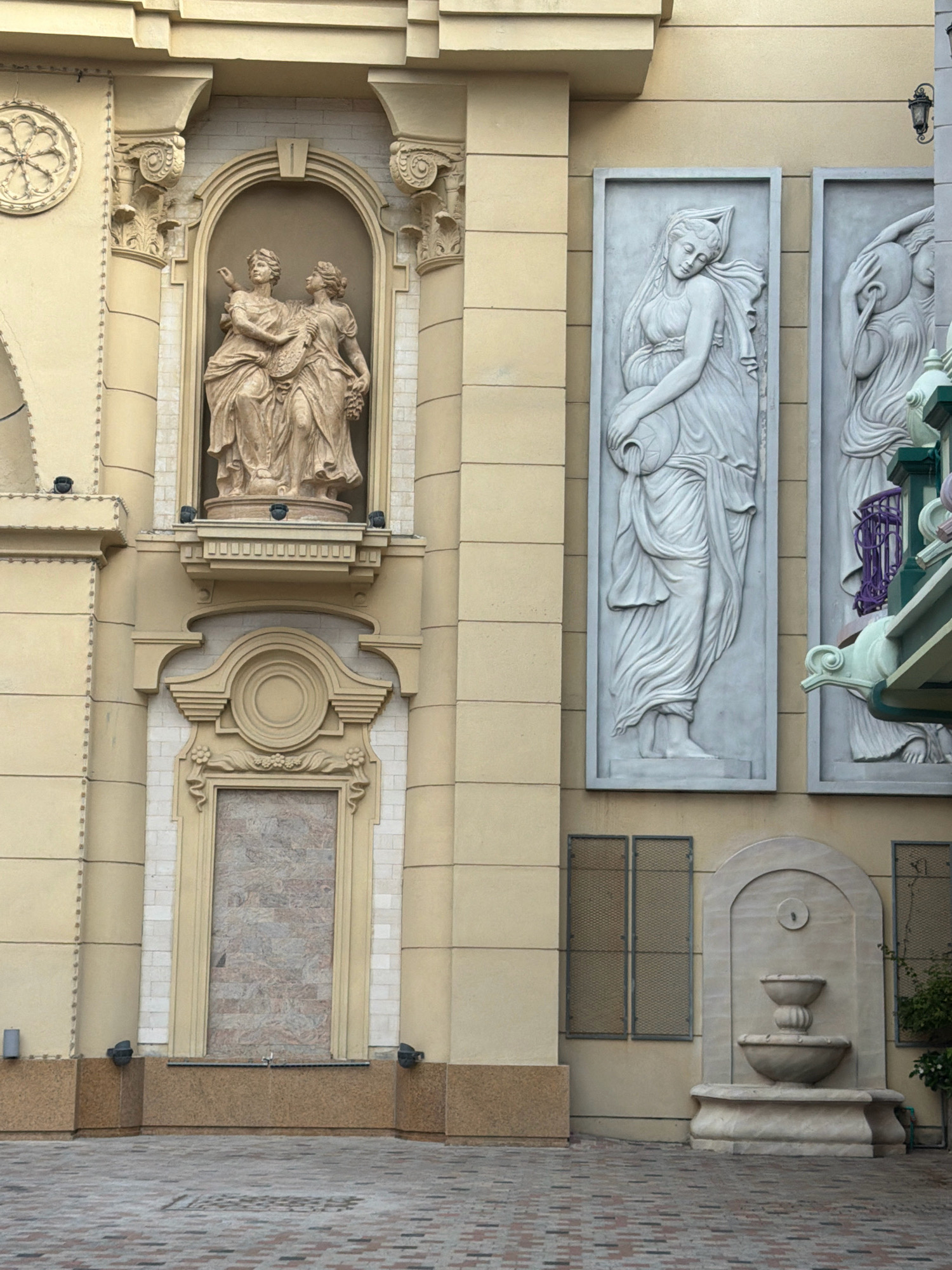 The image shows the exterior of a building with classical architectural elements, likely a hotel or a public building in Nha Trang, Vietnam. The facade is decorated with sculptures and reliefs. On the left, there is an alcove with a sculpture of two draped female figures, one holding what appears to be a lyre and the other a bunch of grapes. Above this alcove is a circular decorative element. To the right of this are two tall, rectangular reliefs depicting draped female figures. The left relief shows a woman holding a large jug from which water appears to be pouring. The right relief shows a woman holding a round object. Further to the right, there is a fountain built into the wall, with multiple tiers and a spout. The building's exterior is painted a light yellow, and the decorative elements are rendered in stone or a material resembling stone. The ground in front of the building is paved with interlocking bricks. The overall style suggests a European neoclassical influence.