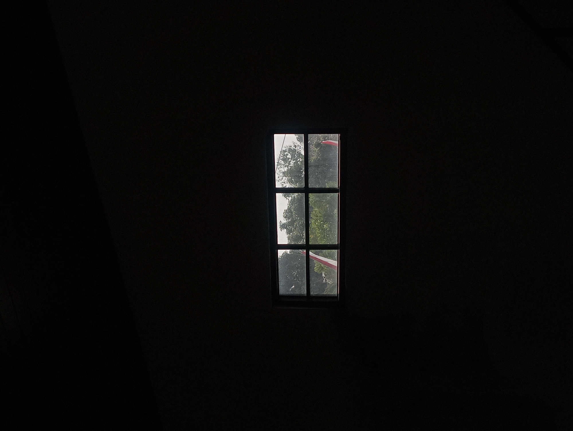 The image depicts a dark, indoor scene with a small, square window in the center. The window is divided into six panes by dark mullions. Through the window, a glimpse of the outdoors can be seen, featuring green foliage and what appears to be an Indonesian flag with red and white stripes. The overall mood is somber and quiet, with the darkness of the interior contrasting with the slightly brighter, overcast scene outside. The location is identified as Surakarta, Indonesia.