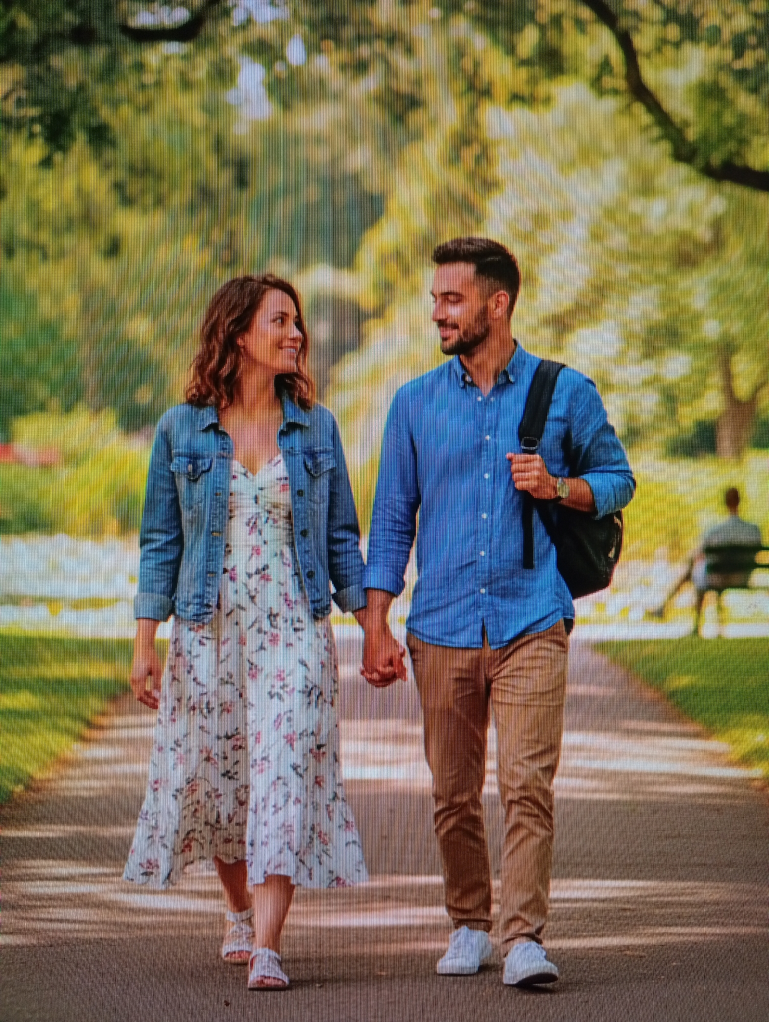 A man and a woman are walking hand-in-hand on a paved path in a park, holding eye contact and smiling. The woman is wearing a floral print dress and a denim jacket. The man is wearing a blue button-down shirt and khaki pants, with a black backpack slung over his shoulder. The park setting features lush green trees and dappled sunlight. In the background, another person is seated on a bench.