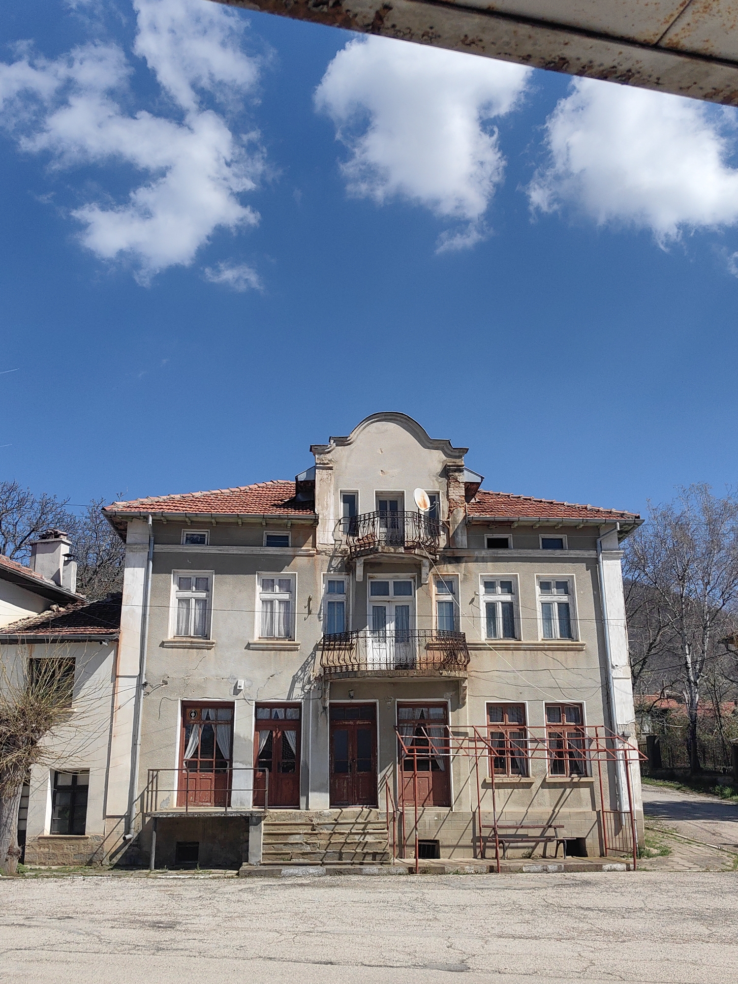 The image captures a three-story building in the village of Hirevo, Bulgaria. The building features a light gray facade, a red tile roof, and multiple windows with white frames. The ground floor has two sets of double doors with dark red frames. Each set of doors is accessible via a short flight of steps. There are visible curtains in some windows. A metal railing surrounds a small porch area on the ground floor. Balconies with ornate metal railings adorn the second and third floors of the building. The sky above is blue with scattered white clouds. A section of rusted metal frame is visible at the top of the image, partially obscuring the sky. Bare trees and foliage can be seen on either side of the building. The ground in front of the building is paved. The overall impression is of a building that has seen better days, with some signs of disrepair.
