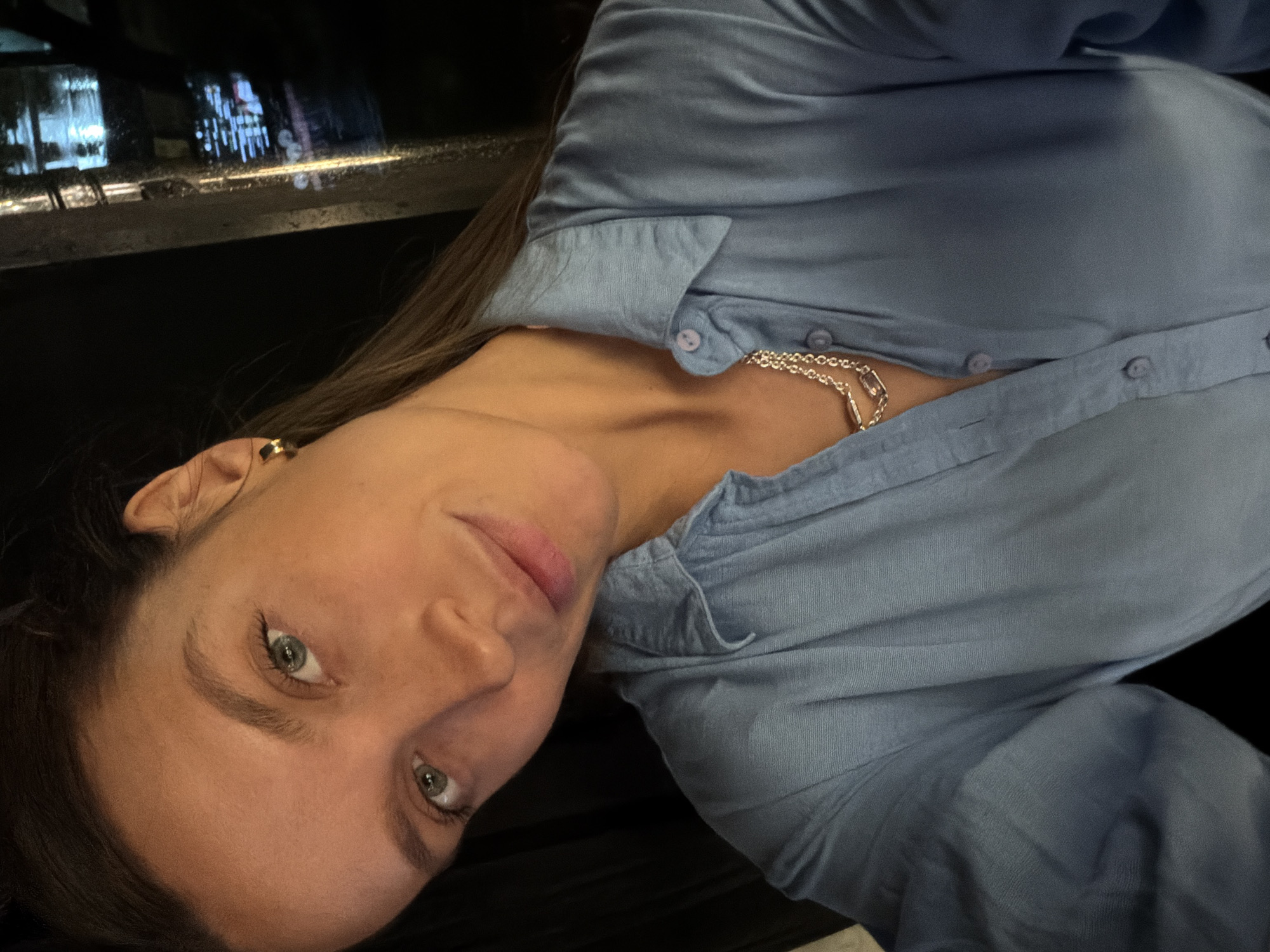 The image captures a person, likely a woman, in an indoor setting during nighttime. The original file is rotated 90 degrees counter-clockwise. The subject is looking directly at the viewer with a calm, subtle smile. She has long dark hair, green eyes, and natural-looking makeup, including pink lips.
She is dressed in a light blue, collared, long-sleeved button-down shirt, with several buttons visible down the front. A silver-colored chain necklace is visible around her neck, and a small gold-toned hoop or stud earring is worn in her left ear.
The background, largely dark, reveals blurred reflections of bright, illuminated city lights from urban buildings through what appears to be a window or glass partition, indicating an outdoor cityscape at night. A dark, smooth surface, possibly a table or bar top, is visible in the immediate foreground. The lighting is focused on the person's face, creating a soft glow against the darker, ambient background, hinting at a cozy evening atmosphere, potentially in Budva, Montenegro, as suggested by the provided context. There is no visible text in the image.