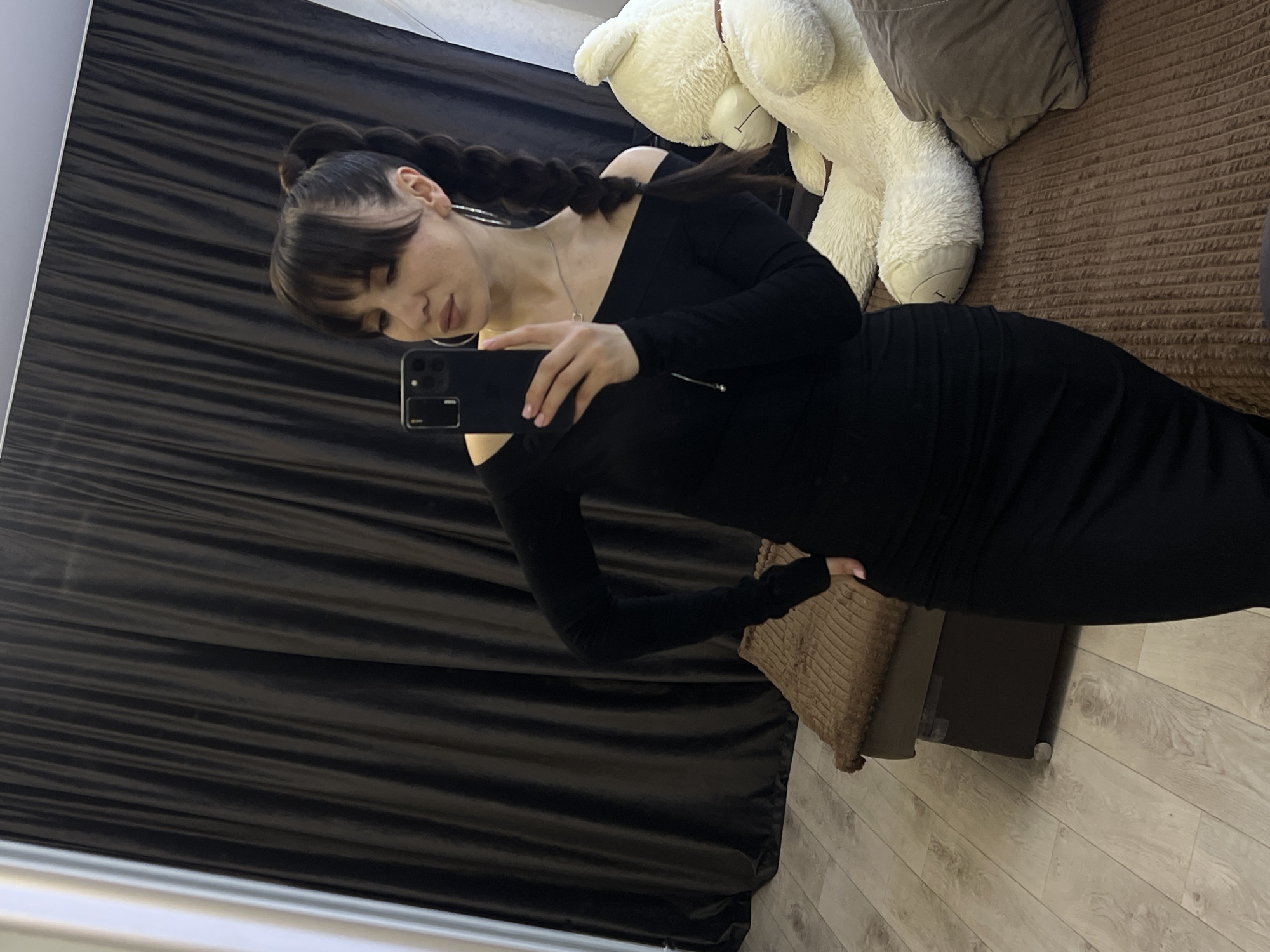 A woman with bangs and long braids is taking a selfie in what appears to be a domestic setting. She is wearing a long-sleeved black dress with a cold-shoulder design and holding a black smartphone up to take the picture. In the background, a white teddy bear and a brown pillow are visible on what seems to be a couch. A dark brown curtain is also in the background, along with a light-colored wood-look floor. It is not possible to determine the specific location in Votkinsk, Russia from the provided image.