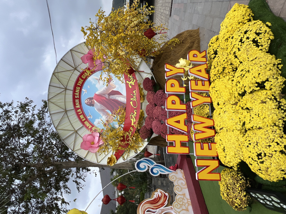 The image displays an elaborate outdoor festive and religious arrangement located at Nha Trang Cathedral (Christ the King Cathedral) in Nha Trang, Vietnam. The scene is captured under an overcast sky.
The central element is a large, circular, light-colored panel featuring a prominent image of Jesus Christ with outstretched arms, set against a blue sky with white doves. Above the image, Vietnamese text reads "ANH EM LÀ ÁNH SÁNG THẾ GIAN" (YOU ARE THE LIGHT OF THE WORLD). Below the image, additional Vietnamese text reads "CHÀO ĐÓN CHÚA XUÂN" (WELCOME SPRING / WELCOME THE SPRING LORD). This circular panel is decorated with artificial yellow blossoms, red ribbons, and two large pink floral elements on its sides.
In the foreground, large, yellow-and-white block letters spell "HAPPY NEW YEAR." These letters are situated amidst a dense, uniform arrangement of bright yellow chrysanthemum-like flowers that extend across the lower part of the frame. Behind the "HAPPY NEW YEAR" sign and beneath the circular Jesus display are multiple mounds of dark reddish-purple flowers and two conical structures covered in a straw-like material. Two red, multi-tiered lantern-like ornaments hang from faux branches on the right side of the display.
To the far left, a partial view of a decorative element with white and blue swirling patterns and a stylized red/orange motif is visible. In the background, a large tree with dark foliage stands to the left of the primary display. Behind the display, the upper section of a large, dark-roofed stone or concrete church building is visible, consistent with the architecture of Nha Trang Cathedral. The ground surface is paved. No individuals are present within the frame.