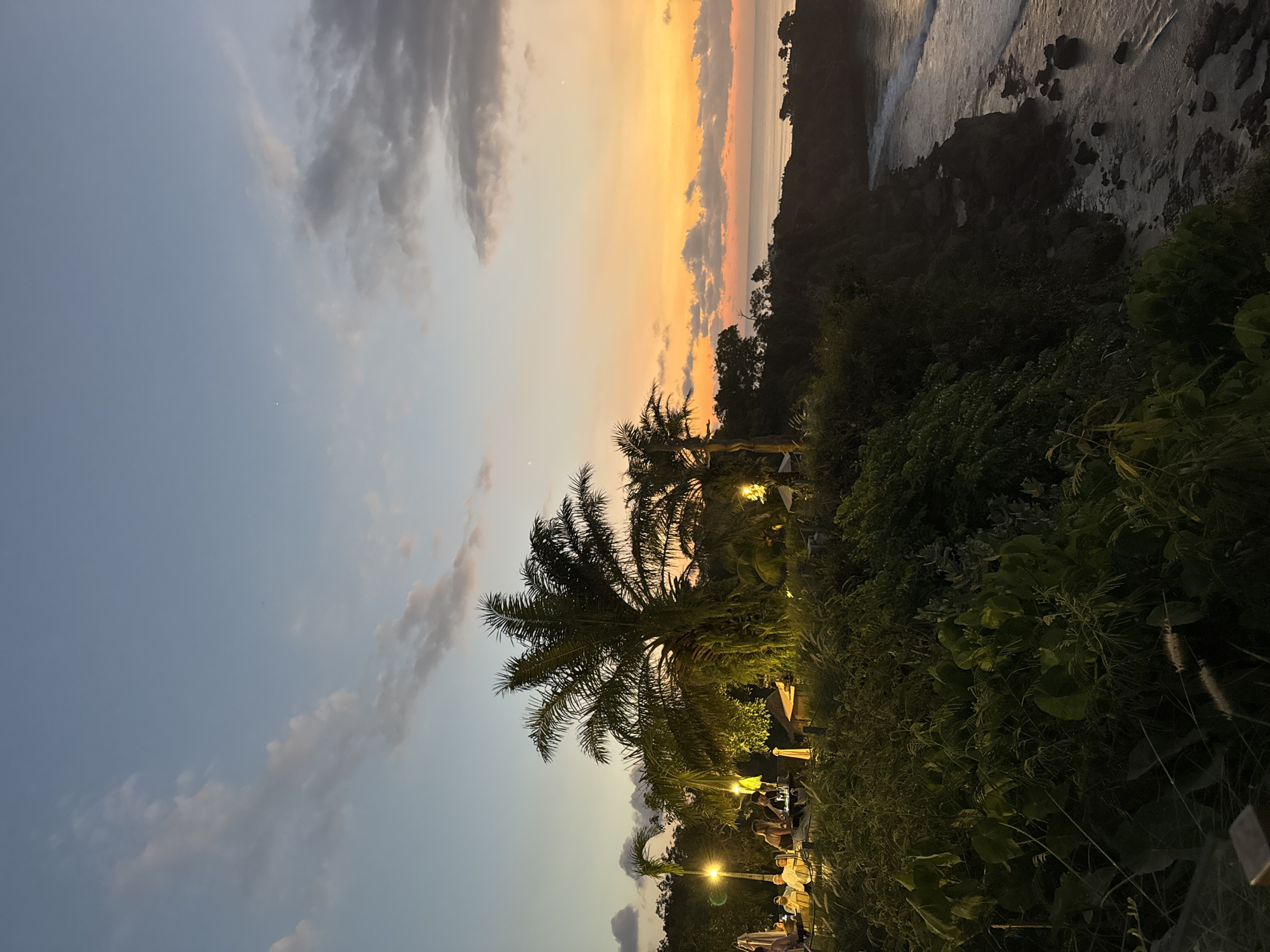 This media file captures a picturesque tropical sunset over a coastal landscape, oriented horizontally for a natural viewing experience. The sky dominates the upper portion of the frame, showcasing a beautiful gradient from a soft blue with scattered white clouds on the left, transitioning into vibrant hues of orange, yellow, and soft pink where the sun dips towards the horizon on the right.
In the midground and foreground, lush green vegetation is abundant. Several tall palm trees are prominent, their dark silhouettes softened by the warm glow of artificial lights positioned beneath and around them. These lights illuminate various parts of the foliage and reveal an outdoor setting where people are gathered. Indistinct figures can be seen sitting or standing, suggesting a social event, a restaurant, or a bar area, enjoying the evening.
To the far right, the dark, rugged outline of a landmass or cliff meets the tranquil ocean. The water surface reflects the warm colors of the sunset, and gentle waves are visible along the rocky shoreline. The combination of natural sunset light and artificial warm lighting creates a serene and inviting atmosphere.
The time of day is clearly dusk or sunset. The weather appears calm and clear, with only light, scattered clouds. The presence of palm trees, the ocean, and lush vegetation strongly indicates a tropical or subtropical coastal location. No visible text is present in the image.