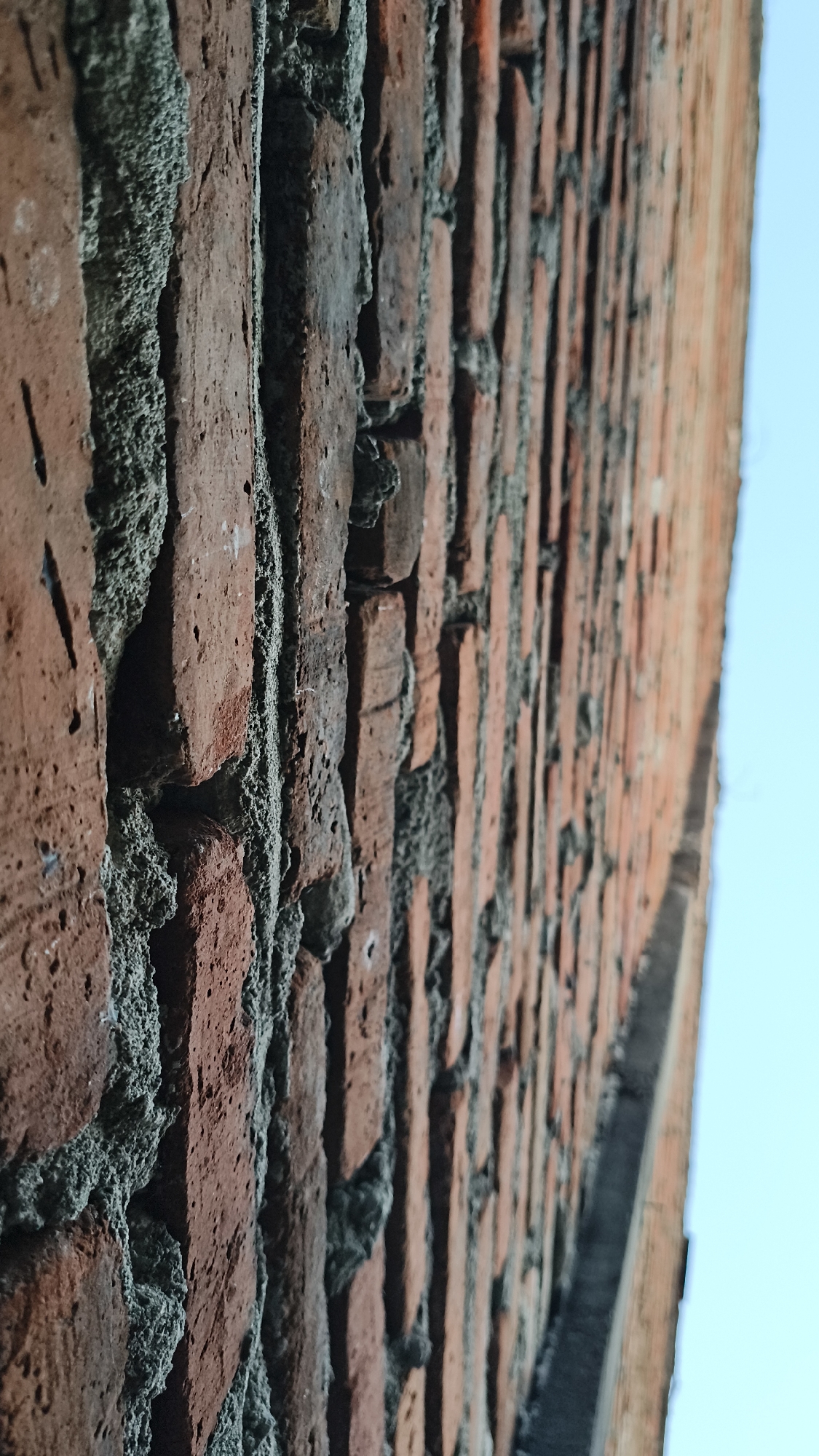 The image shows a close-up, side-view of a brick wall with visible mortar. The bricks are reddish-brown and vary in texture, with some appearing rough and chipped. The mortar is gray and irregularly applied, creating a textured surface between the bricks. The wall extends from the bottom left to the top right of the frame, with the perspective leading the eye further down the wall. To the right of the wall, a clear, light blue sky is visible. The scene is static and offers no information about people, actions, or interactions. This image does not contain any elements that would allow for identification of its location in Kerep Kidul, Indonesia.