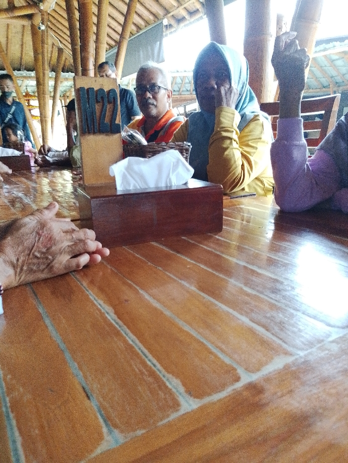 The image depicts an indoor scene at a restaurant or cafe in Pabelan, Indonesia. The foreground features a light brown wooden table with visible grains and light reflections. On the table, from left to right, are a partially visible human hand with prominent veins, a dark brown wooden tissue box containing white tissues, and a light brown wooden table number sign displaying "M22" in black lettering.
In the midground, several individuals are seated. A man with short grey hair and glasses, wearing an orange and dark blue collared shirt, looks directly at the camera. To his right, a woman in a light blue hijab and a yellow long-sleeved shirt rests her right hand on her cheek, looking away from the camera. Further to her right, a partial arm in a purple long-sleeved garment is visible, resting on the table. In the background, other indistinct figures, including one man wearing a dark shirt and a face mask, are present.
The environment includes visible bamboo support columns and ceiling beams, suggesting an open-air or semi-enclosed structure. Bright natural light illuminates the scene from an unseen source, likely an open side or window. The overall setting is a casual dining area.