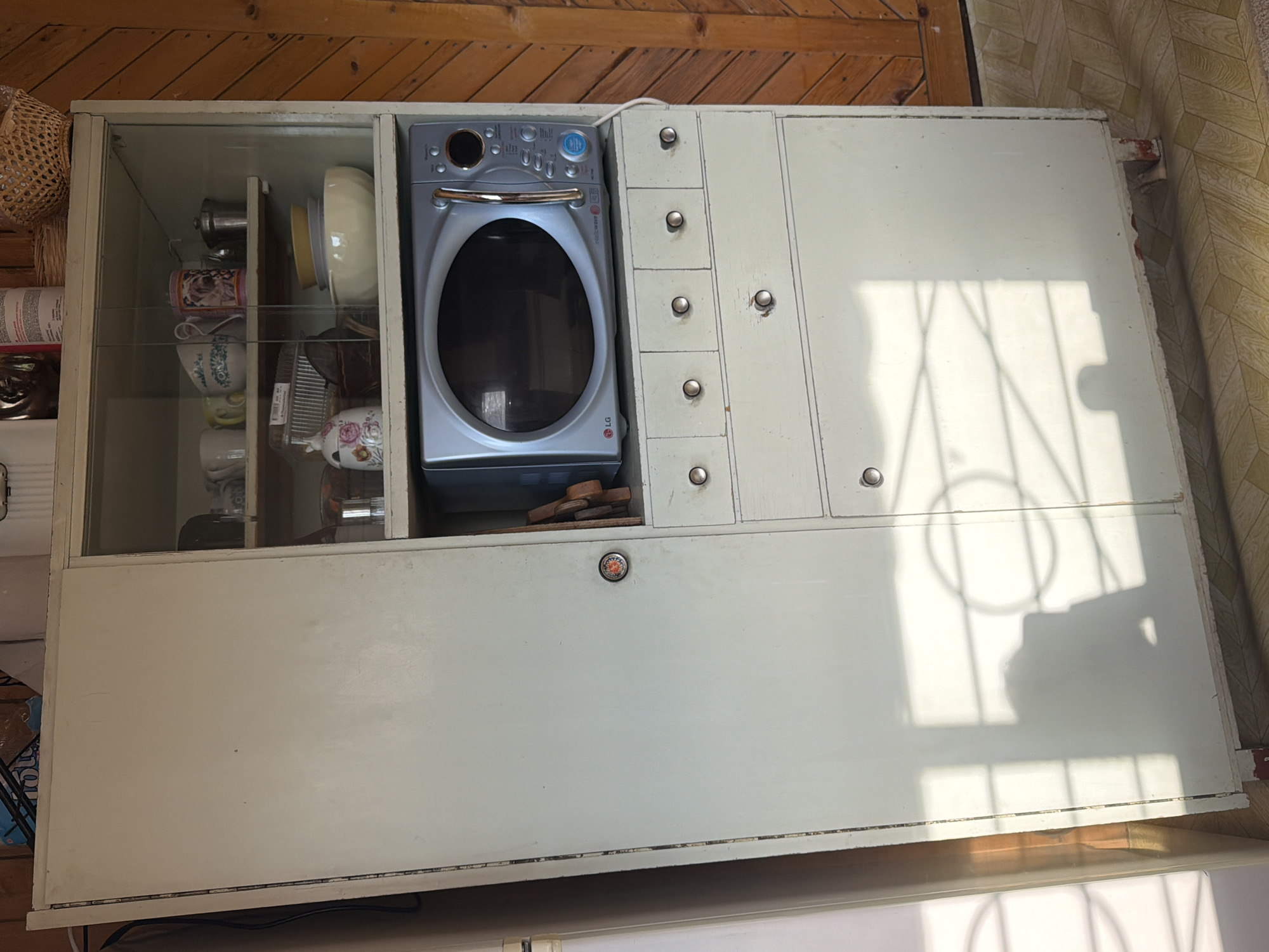 A high-angle shot displays a pale white kitchen cabinet. The cabinet is divided into sections. The leftmost section is glass-doored, revealing a collection of dishes and kitchenware. To the right, an LG washing machine is encased in the cabinetry. Next to the washing machine, there are six small drawers with silver knobs, positioned vertically. The rightmost section of the cabinet appears to have two doors and a floral-patterned knob. The cabinet sits on a wooden floor near patterned wallpaper. Based on the prompt information, this scene is located in Plamya, Russia.