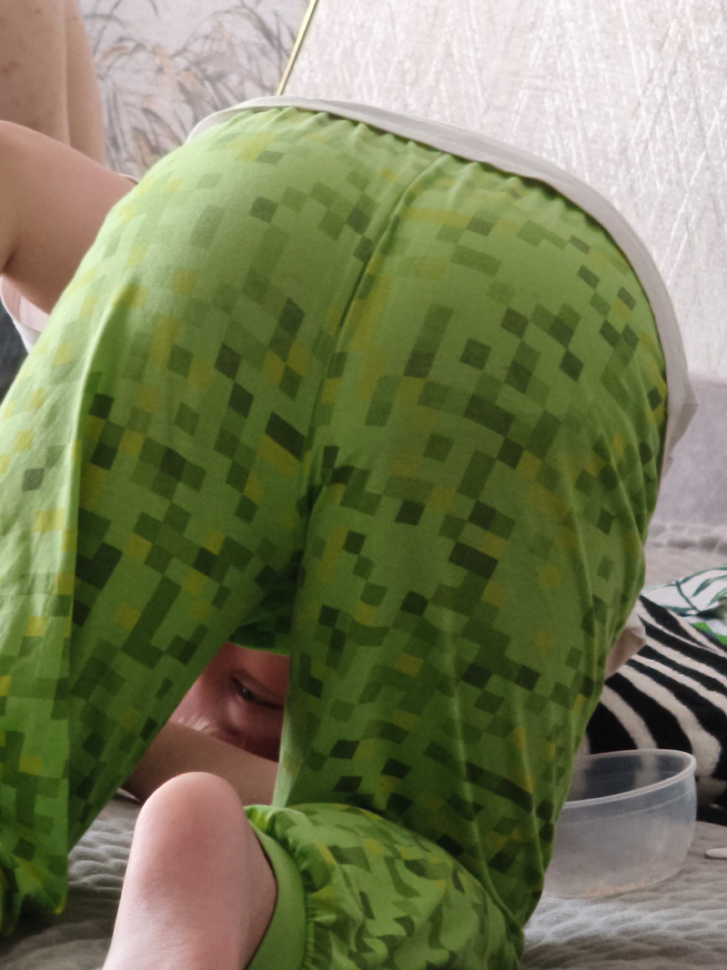 This is a medium shot, slightly high-angle photograph of a person wearing green pixelated pajamas. The person is bent over, their back is to the camera, and their head is tucked between their legs, peeking out from underneath the pajamas. Their face is partially visible, with one eye looking towards the camera. The pajamas have a pixelated pattern of varying shades of green and yellow. The background is blurred but appears to be a domestic setting, possibly a bedroom or living room, with a textured surface that could be a bed or couch. There is a white object, possibly a piece of clothing or bedding, visible behind the person's left shoulder. To the right, a portion of a zebra-striped pillow and a clear plastic bowl are visible. The lighting suggests it is daytime, likely indoors. The scene has a playful and casual feel. No text is visible in the image. The location context is Chaikovsky, Russia.