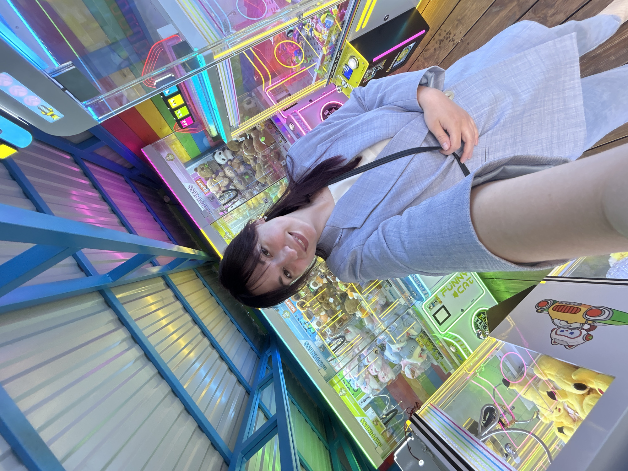 A woman in a light blue blazer takes a selfie in front of a row of claw machines filled with stuffed animals. The background features a colorful rainbow wall and a metal roof supported by blue beams. The claw machines have bright neon lights and are filled with various plush toys, including bears, pandas, and sharks. The woman is smiling and holding her phone with her right hand. The overall scene appears to be an amusement or entertainment venue.