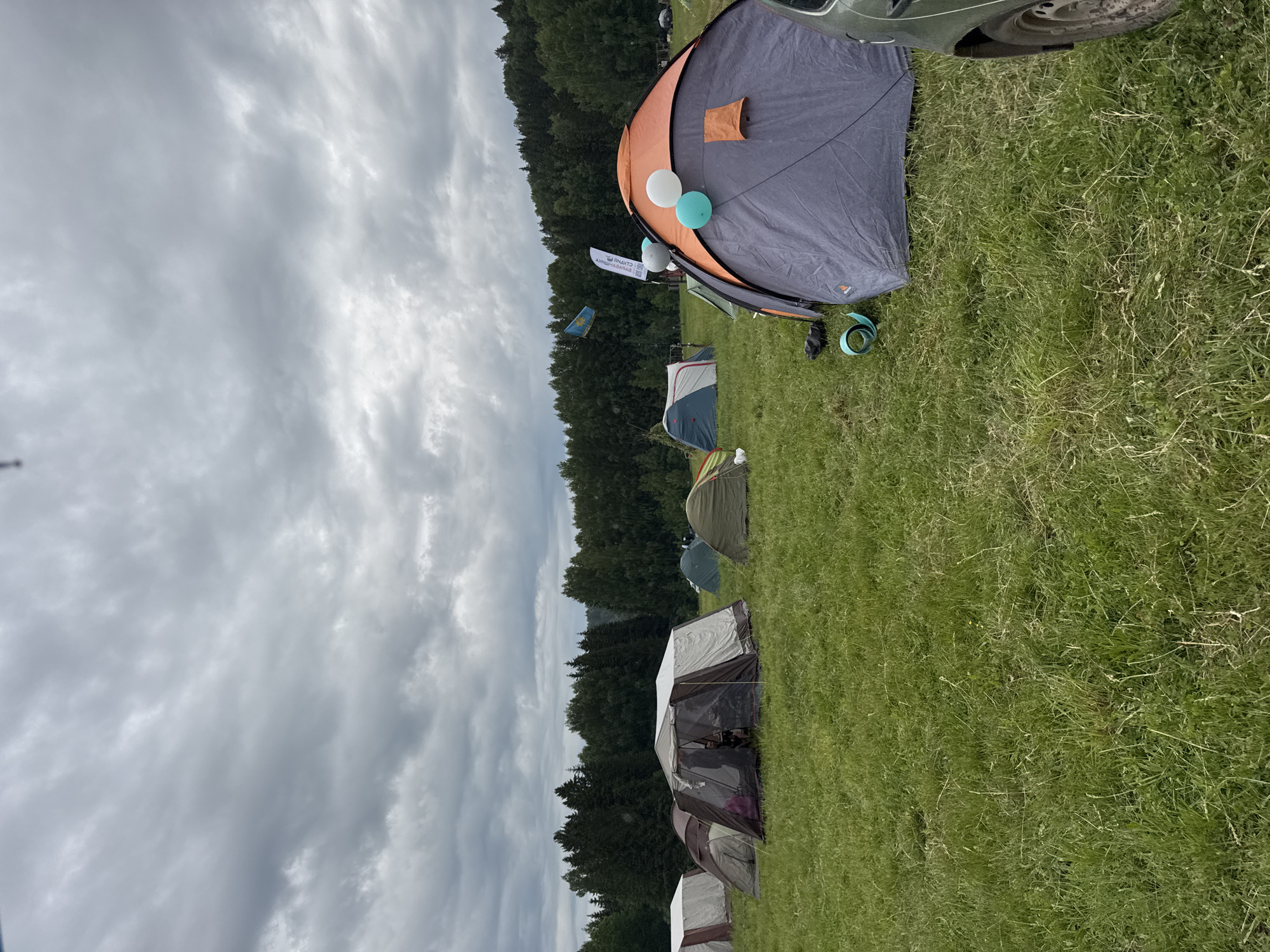 This image captures an expansive outdoor camping scene in Bol'shaya Kivara, Russia, under an overcast sky. The setting is a large grassy field, bordered by a dense line of dark green conifer trees that form a forest in the background.
Numerous tents of various sizes and designs are pitched across the field. In the foreground, a prominent grey and orange dome tent is partially open, adorned with two balloons—one white and one light blue/turquoise—near its entrance. A small, light brown pouch is attached to the side of this tent. To its right, the front wheel and part of the bumper of a dark-colored car (possibly green or grey) are visible. On the grass near this tent, a turquoise strap or band and a dark object are present.
Further into the campsite, a line of other tents can be seen, varying in color from green to grey and white. These include dome-style tents and potentially larger cabin tents. No people are explicitly visible in the frame, but the numerous tents imply a significant gathering.
An activity or event appears to be taking place, given the clustered tents and decorative elements like balloons and flags. Two flags or banners are visible on poles: a white banner near the foreground tent features text in Cyrillic script, which appears to read "СТАНАЯ САРАБА" (STANAYA SARABA) when viewed upright. Further back, a blue flag with a yellow design or emblem is flying.
The time of day is daytime, but the weather is characterized by a heavily clouded, grey sky, suggesting either a recent rainfall, high humidity, or impending precipitation. The overall atmosphere is functional, set for an outdoor gathering, with elements of festivity indicated by the balloons.