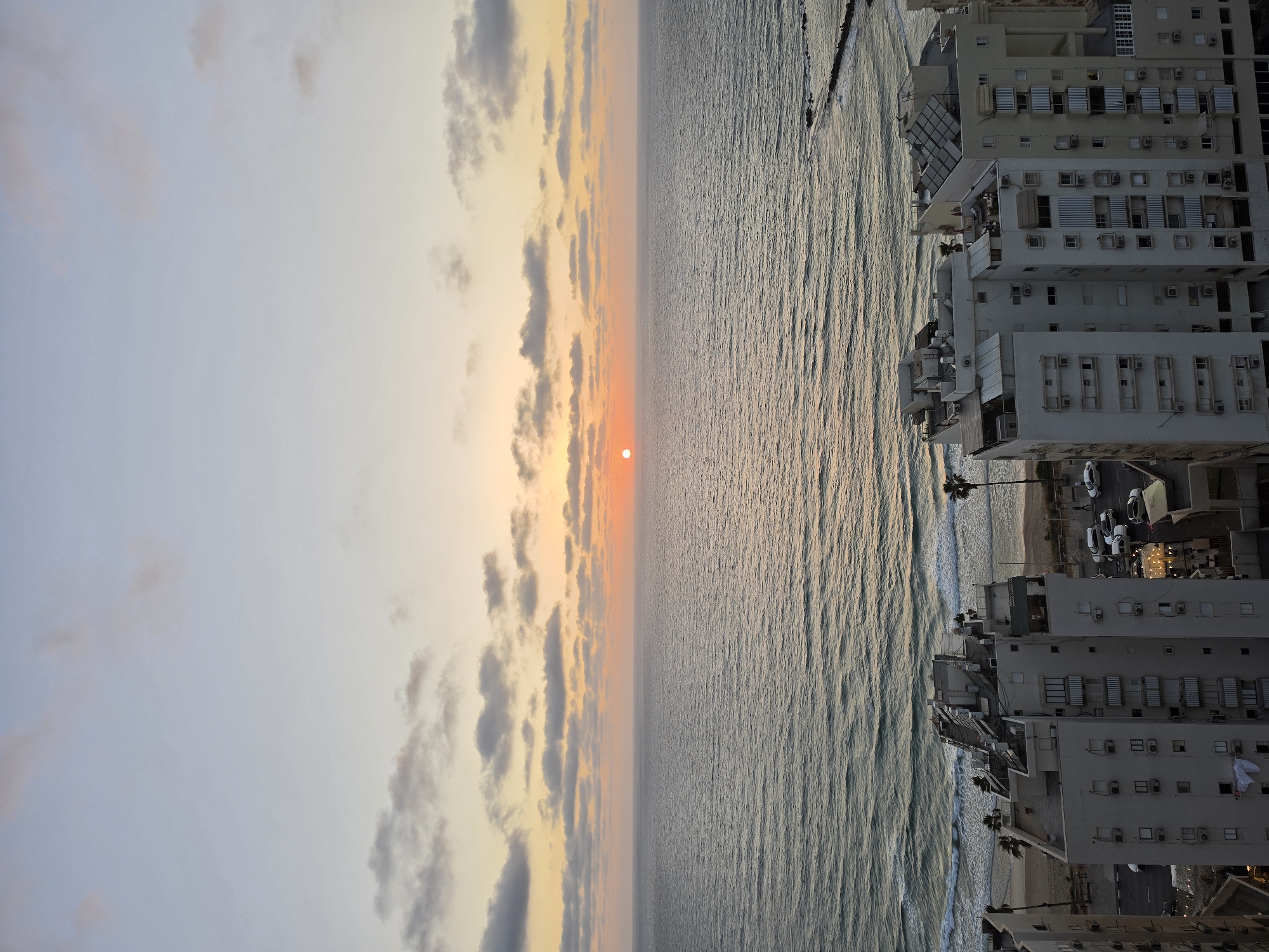 This image captures a vibrant sunset over the coast of Bat Yam, Israel. The scene is divided horizontally into three main sections: the sky, the sea, and a coastal city with a beach.
In the upper portion of the frame, the sky displays a gradient of colors, transitioning from a soft, pale blue at the top to warm orange and yellow hues near the horizon. The setting sun, a prominent orange-red orb, is visible low on the horizon, partially obscured by light haze or clouds. Scattered clouds, some dark grey and others tinged with orange from the sunset, are visible across the sky.
The middle section features the vast expanse of the sea, with gentle waves reflecting the golden light of the setting sun. The water's surface shows a textured pattern of ripples and waves extending towards the horizon.
The lower portion of the image reveals a densely built coastal area. Numerous multi-story buildings, predominantly light-colored, line the coastline. These structures appear to be residential or hotel buildings, characterized by multiple windows, balconies, and visible air conditioning units. A strip of sandy beach separates the buildings from the sea, and several palm trees are visible along this beachfront. Below the buildings, what appears to be a street or parking area is visible, with several cars. Some artificial lights are already illuminated on the buildings and in the city area, indicating that it is dusk or early evening.
The overall impression is a peaceful and scenic view of a coastal city at sunset, blending natural beauty with urban architecture. There are no prominent human figures or specific activities visible, and no text is discernible within the image.