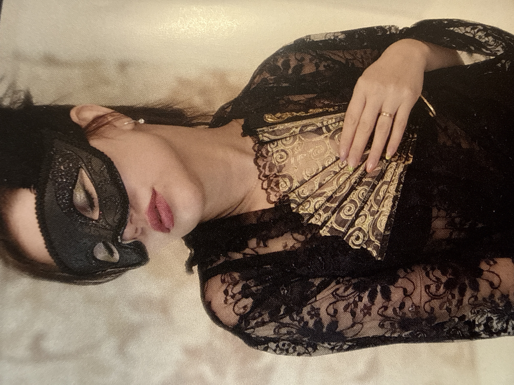 A woman wearing a black sequined mask and a black lace robe holds a fan in front of her chest. She has dark hair and is wearing lipstick. A thin gold ring is visible on her right ring finger. The background is a textured, light-colored wall. The image is a close-up shot, focusing on the woman's face, shoulders, and the fan. There is no identifiable location in the city of Cheboksary, Russia.