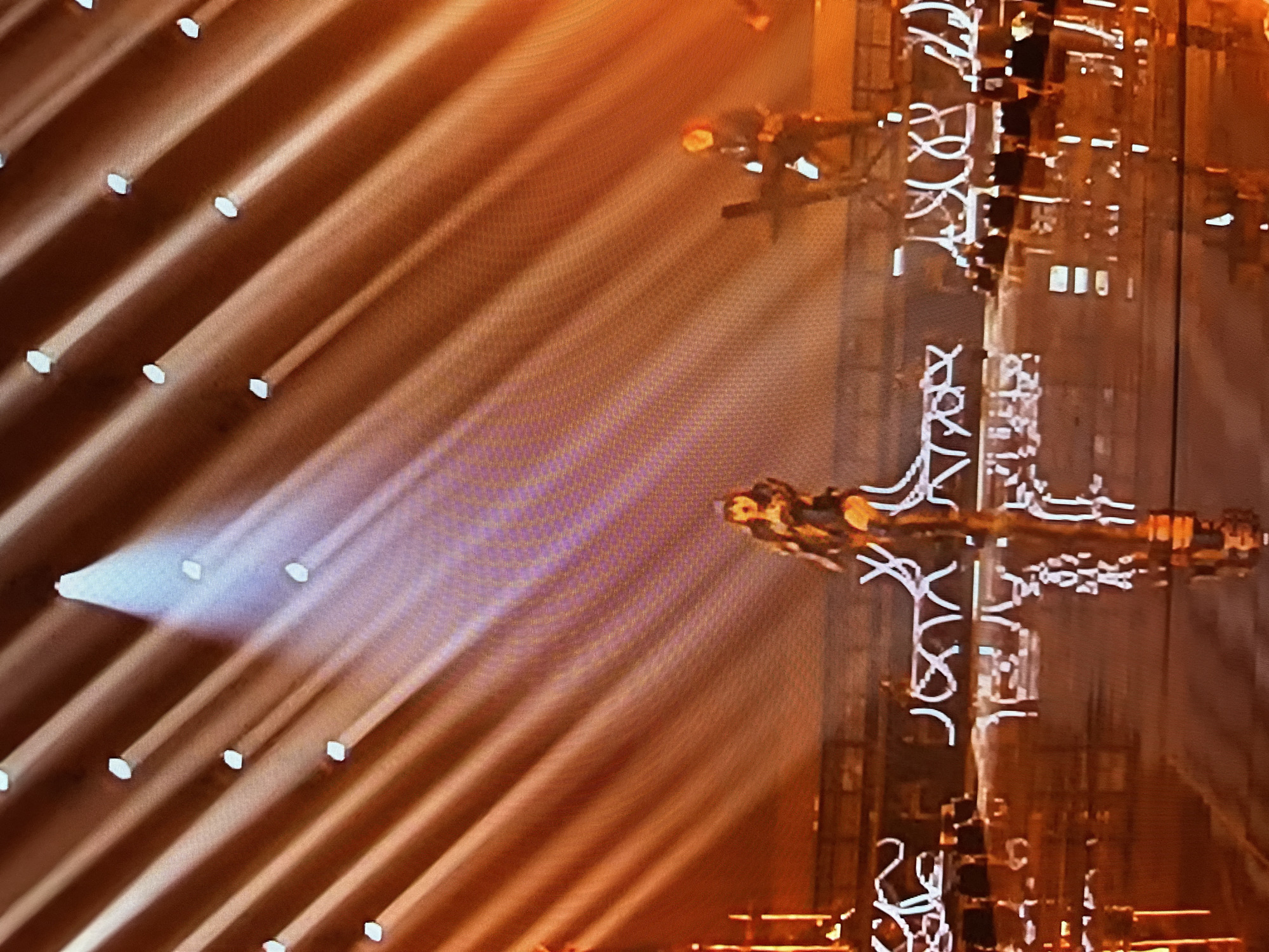 The image shows a vertical structure adorned with lights and what appear to be decorative elements, potentially stylized Cyrillic script. Two figures in similar poses are positioned on either side of the structure. The lighting is dramatic, with beams shining from the left, illuminating the figures and the structure. The scene appears to be a performance or staged event, with a warm color palette dominating the image. Without additional information, specifying a precise location in Sofia, Bulgaria is impossible.
