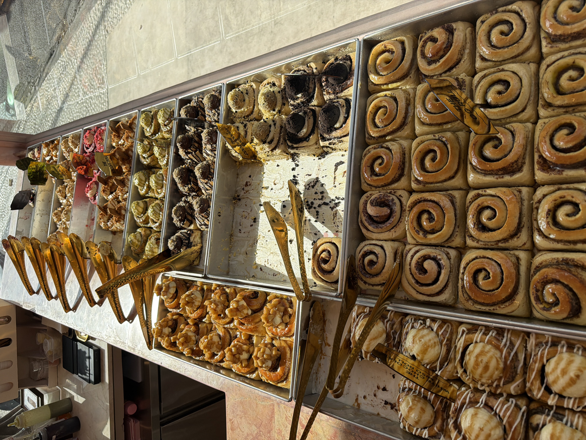 The image shows a display of various baked goods, likely cinnamon rolls, arranged in metal trays on a counter. The trays are stacked and tilted, with golden tongs positioned above them, suggesting they are available for purchase. The baked goods vary in toppings and presentation, with some appearing to be plain, while others are adorned with drizzles of icing, nuts, or fruit. In the background, bottles of sauces and a scale are visible on a counter, as well as a window with light filtering through, casting shadows on the display. The overall scene suggests a bakery or pastry shop. The specific location in North Kuta, Indonesia is not explicitly indicated by any visual cues in the image.