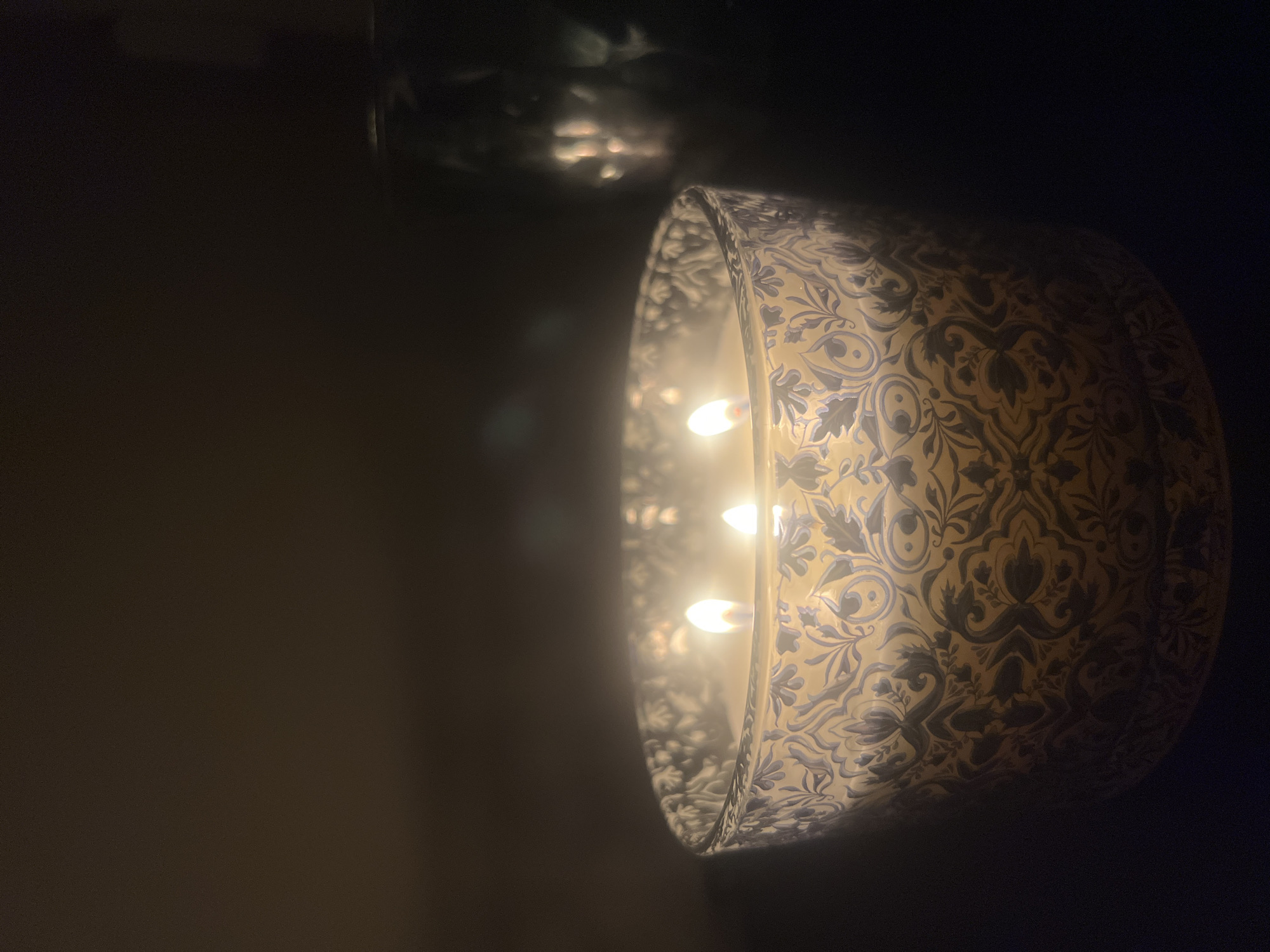 A lit candle with three wicks sits on a dark surface. The candle is in a patterned container with a light tan or yellow background and dark blue designs. The dark background suggests it is nighttime or a dimly lit room. There is a blurred reflection of light in the upper left corner, possibly a reflection of the candle flame. The photo is taken at a slightly high angle, with the candle being the main subject, occupying the right side of the frame. There is nothing in the image to suggest it is in Sofia, Bulgaria.