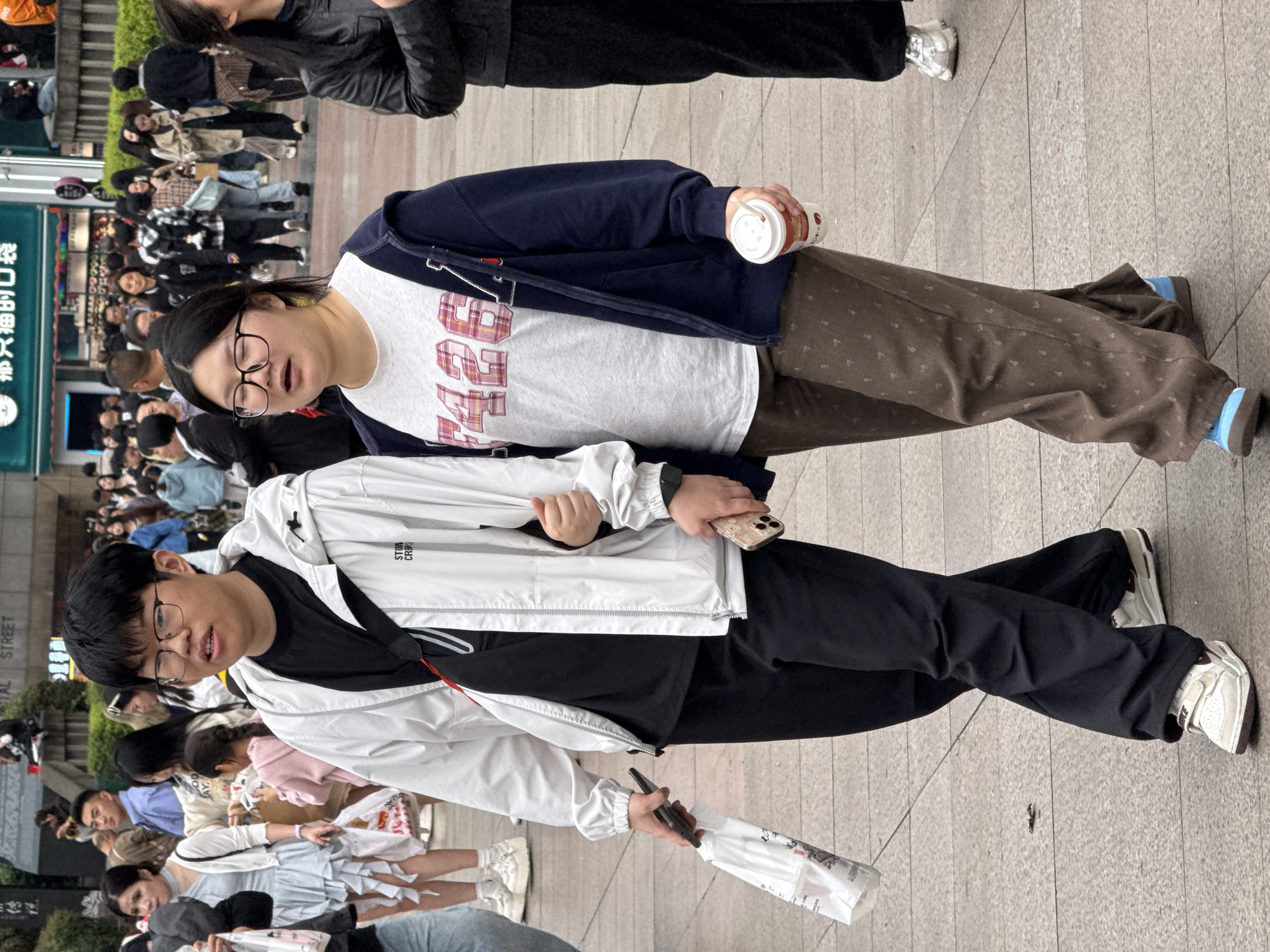 Two people stand in the foreground on a gray paved surface. The person on the left wears glasses, a black t-shirt, a white jacket, and black pants, holding a phone and a white bag. The person on the right wears glasses, a white t-shirt with a large graphic, a navy blue jacket, and patterned brown pants, holding a cup. In the background, a crowd of people stands near a storefront with Chinese writing, suggesting a location in China. The name "CAPITAL STREET" is visible on a sign. The location is likely in Liangjiang, China.