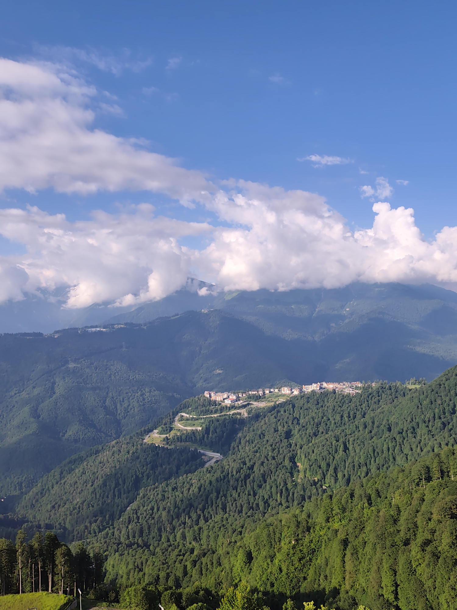 The image captures a panoramic view of a mountainous landscape in Estosadok, Russia, under a bright blue sky dotted with fluffy white clouds. In the foreground, a dense forest of tall, green trees covers the slopes of the mountains. A winding road snakes through the trees, leading up the mountainside.
Further up, nestled on the side of a mountain, is a cluster of buildings, appearing to be a resort or residential area. These structures are modern, with light-colored facades and multiple stories, suggesting they are hotels or apartment complexes. A road is visible leading to and from this settlement, winding its way through the terrain.
In the distance, larger, more imposing mountains rise, their peaks partially obscured by clouds. The overall impression is one of natural beauty and human development integrated into a rugged mountain environment. There are no people visible in the image.