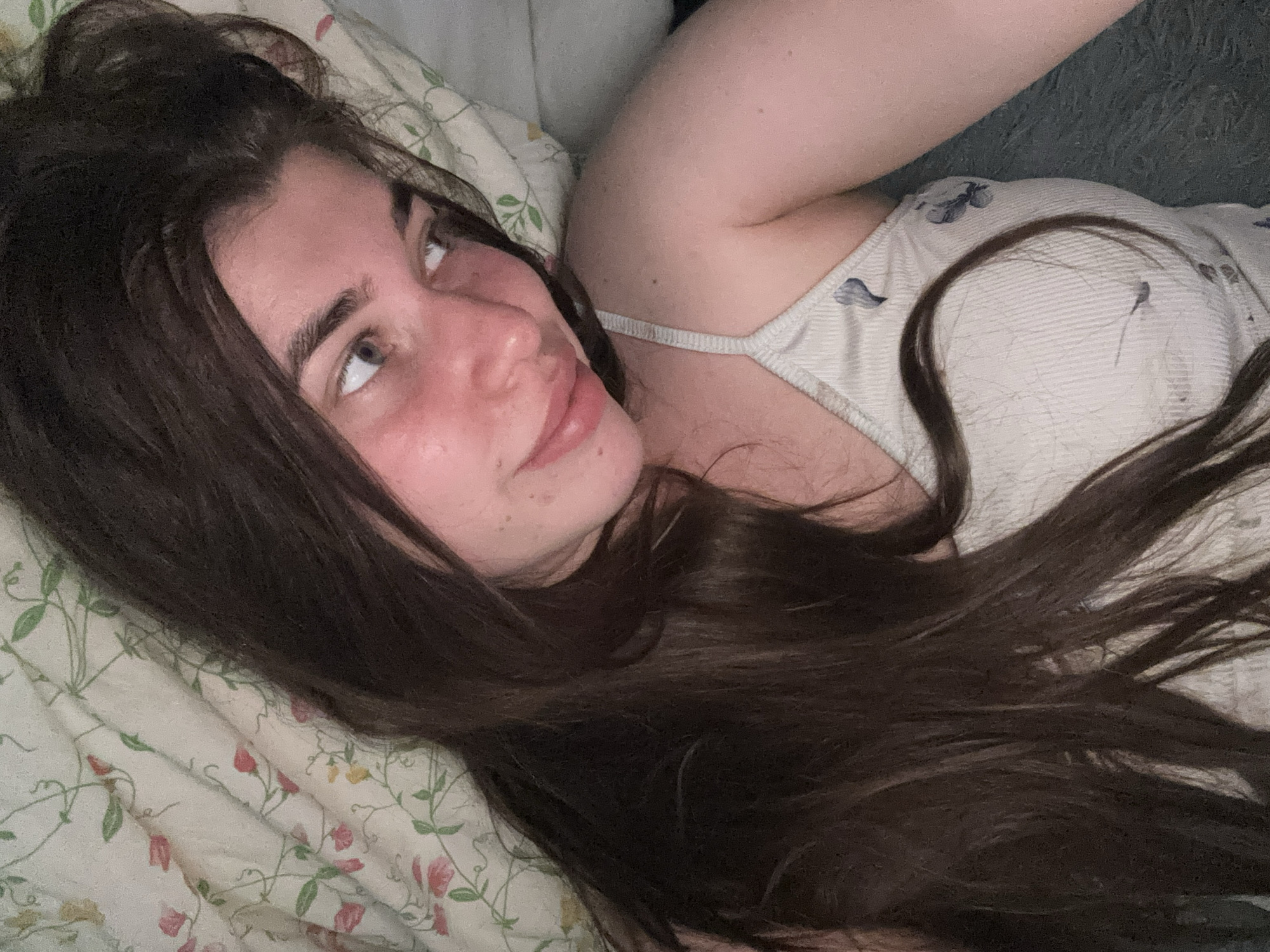 This media file shows a close-up, overhead shot of a young woman lying down in what appears to be a bed. She has long, dark brown hair spread out around her, and she is looking up and to her left. She is wearing a light-colored tank top with a subtle pattern. The bedding beneath her has a floral print with small pink and green flowers. The overall lighting suggests it might be nighttime or a dimly lit indoor setting, possibly morning. Her skin appears flushed, and her expression is relaxed, perhaps contemplative. There are no visible text elements in the image. The location context is Мещёры, Russia, which suggests a potentially rural or natural setting, though the immediate scene is indoors.
