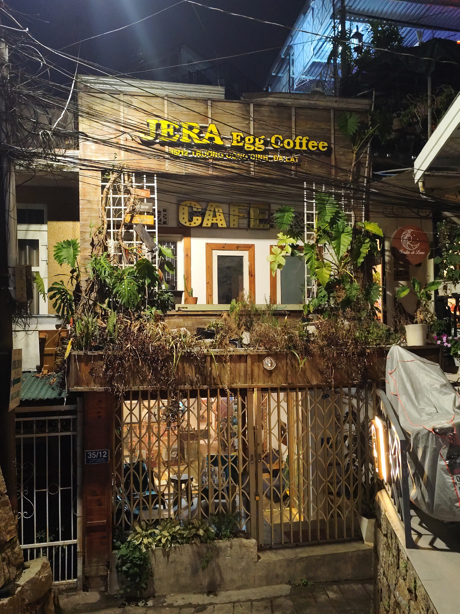 The image displays the illuminated facade of a multi-story building at night, identified as "JERA Egg Coffee" in Da Lat, Vietnam. The building features a rustic design with wooden paneling on its upper sections and a mix of lush green and dry, trailing plants arranged in planters and climbing trellises on the second floor.
A prominent yellow sign spells out "JERA Egg Coffee" across the upper part of the building. Below it, smaller text reads "242 TRƯƠNG ĐỊNH, ĐÀ LẠT," providing the specific address. Beneath these, a large, illuminated sign spells "CAFE." The ground level entrance is secured by an ornate, closed metal gate, through which interior lighting reveals cafe seating. To the left of the main gate, a smaller white gate with an address plaque "35/12" is visible. Overhead, a dense tangle of utility wires crosses the frame against the dark night sky.
On the right side of the image, beyond the cafe entrance, a covered motorbike is parked next to a stone wall. Above the motorbike, a circular sign displays a crescent moon and the text "Jardin de Luna." Several potted plants are placed around the entrance and on the upper ledges. The scene suggests a typical evening in an urban area of Da Lat, with the cafe likely closed or operating behind its secured entrance. No people or specific activities are visible, conveying a static night-time street view.