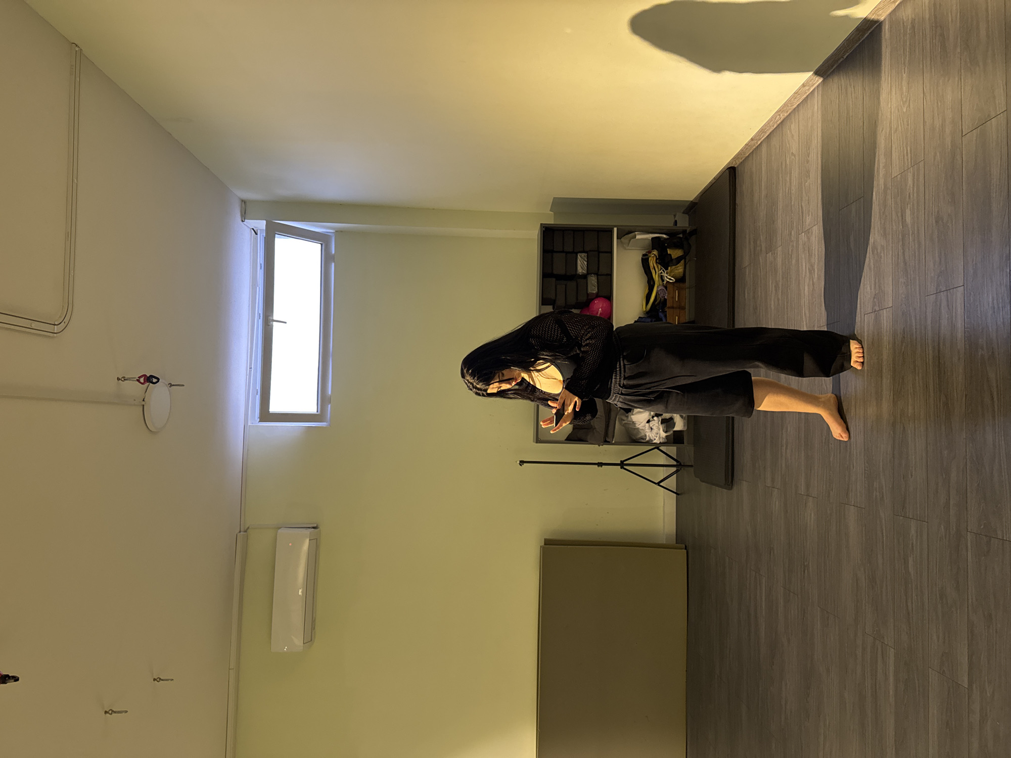 The image shows an interior space with a light green wall and gray wood-patterned flooring. A woman with long black hair is standing barefoot in the room, wearing black clothing. She appears to be in motion, with her hands raised. Behind her is a mirror on a stand, and a shelving unit is visible in the background containing various items. A window is situated on the wall to the left, and an air conditioning unit is mounted below it. Several small fixtures are also visible on the wall. A large, light-colored rectangular panel is standing against the wall further away from the window. The location is Unknown, Unknown.