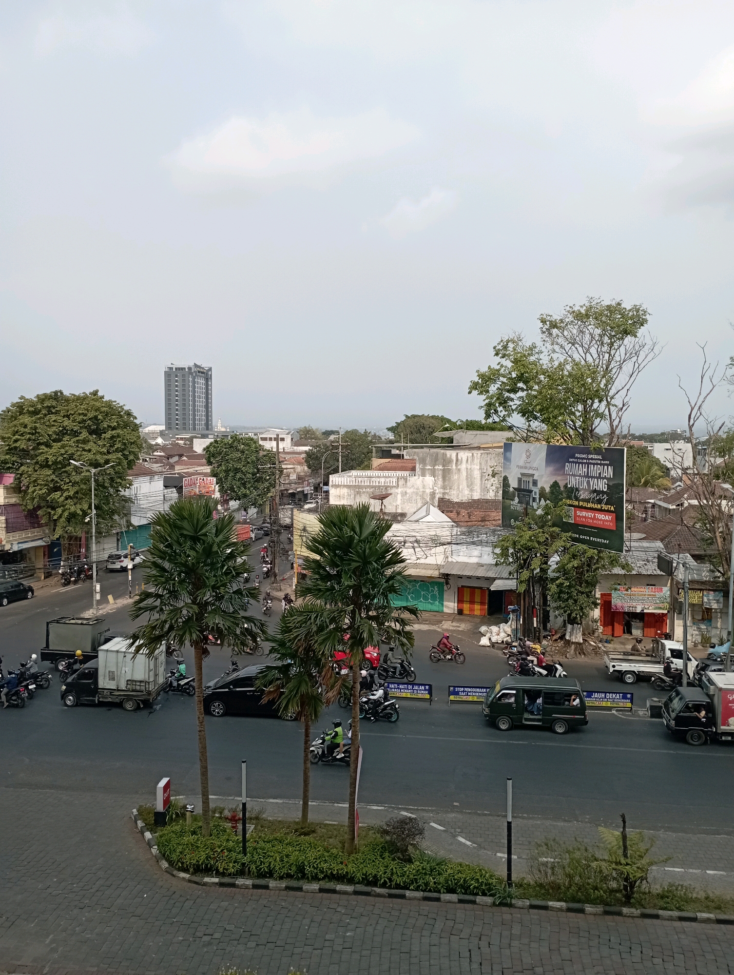 This is a photograph taken from an elevated perspective overlooking a street intersection in Malang, Indonesia, likely at dusk. The scene is characterized by urban traffic and commercial activity.
In the foreground, a paved walkway with a cobble-like pattern curves towards the intersection. A small patch of green landscaping with plants and a fern borders the walkway. A single pole with a red and white indicator labeled "OUT" is visible on the edge of the walkway. Two tall palm trees with segmented trunks stand prominently, their fronds reaching towards the sky.
The intersection itself is busy with vehicles. Multiple motorcycles are present, some parked, others being ridden by individuals wearing helmets. A dark-colored car is positioned at the intersection, and a light-colored truck with an open cargo area is also visible. A dark green minivan is traveling through the intersection, and a black truck with a closed cargo box is further down the street. Several small shops and storefronts line the sides of the road, with some displaying signage. One notable sign is a large billboard advertising a housing development. In the distance, a tall, modern building dominates the skyline, indicating a more developed part of the city. The sky is overcast, with patches of light clouds, suggesting either early morning or late afternoon lighting.