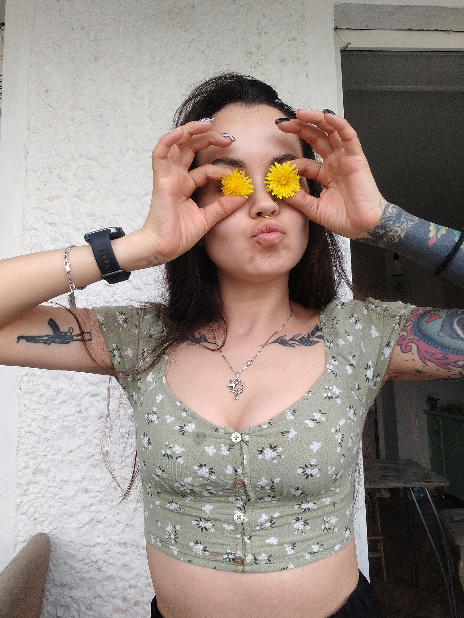 A young woman is posing outdoors, holding yellow dandelions over her eyes as if they are glasses. She has long dark hair, a nose ring, and tattoos on her arms, chest, and neck. She is wearing a cropped floral-patterned top. A black smartwatch and a silver bracelet are visible on her left wrist. The background includes a white wall, a doorway, and a glimpse of the interior of a room. The location is Noviy, Russia.