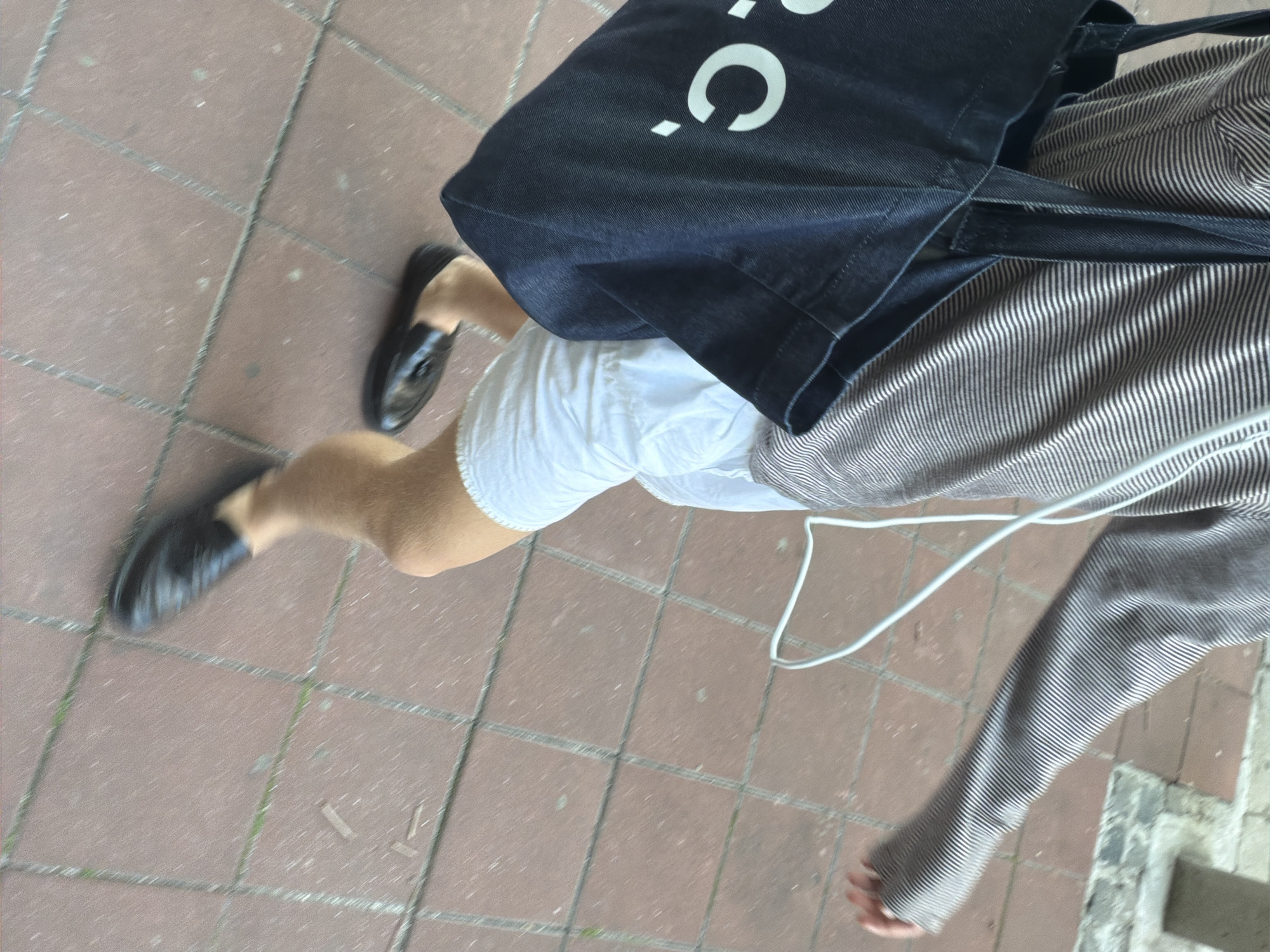 This media file shows a person walking on a sidewalk made of reddish-brown tiles. The person is wearing black loafers, white shorts, and a striped long-sleeved shirt. They are also carrying a dark blue tote bag with a white logo on it. The logo appears to be a stylized 'C' and a dot. A white cord, possibly part of earbuds or a charger, is draped over the person's arm and the tote bag. The image is taken from a high angle, looking down at the person's legs and torso. The lighting suggests it is daytime, and the overall scene appears to be outdoors in an urban environment, consistent with a location in Belgrade, Serbia. The focus is on the person's attire and accessories, with the background showing the textured sidewalk and a glimpse of a concrete edge on the right side of the frame.