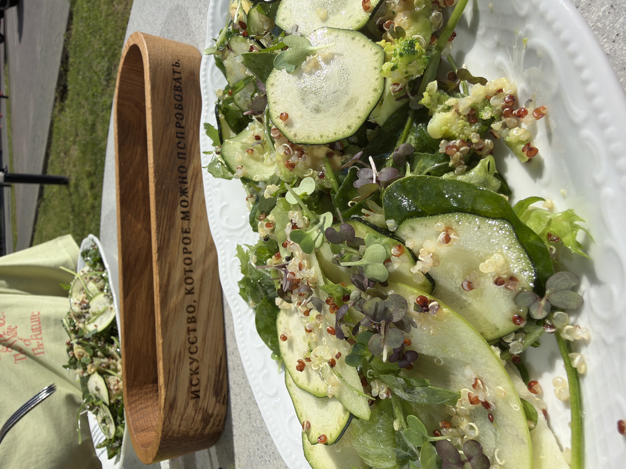A close-up, slightly angled overhead shot shows a white ceramic platter filled with a vibrant salad. The salad is a mix of sliced zucchini, broccoli florets, quinoa, and dark green leafy vegetables, possibly spinach or arugula. Tiny reddish-purple sprouts are scattered throughout, adding a splash of color and texture. The ingredients are glistening, suggesting they are dressed.
To the left of the platter, a wooden salad tong rests on a light green cloth napkin. The tong has a curved, U-shaped design and is engraved with text in Russian. The napkin appears to be part of a table setting, with a fork visible beneath it. In the background, out of focus, is a patch of green grass and a hint of a paved surface, possibly a street or pathway.
The lighting suggests it is daytime, likely outdoors, with natural light illuminating the food. The scene appears to be a casual outdoor dining setting. The visible text on the wooden tong, in Cyrillic script, reads: "ИСКУССТВО, КОТОРОЕ МОЖНО ПОПРОБОВАТЬ." which translates to "Art you can try." This suggests the salad is presented as a culinary creation, akin to art. The overall impression is one of freshness and a focus on healthy, well-prepared food. No people are visible in the frame. The location is identified as Saint Petersburg, Russia.