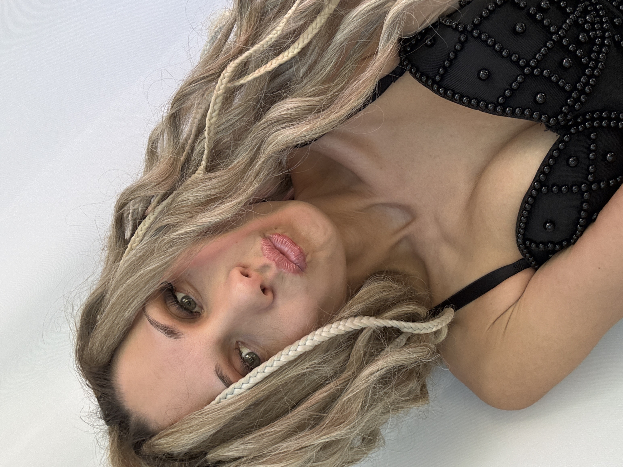An adult female is depicted in a close-up, angled portrait taken in Rusanovo, Russia. Her head is tilted, and she directly faces the camera with a puckered lip expression. Her hair is long, wavy, and multi-toned blonde with visible darker roots, featuring several thin, light-colored braids interwoven throughout the strands, some draped forward over her shoulder and face. Her eyes are green, and she has well-defined eyebrows. She is wearing a dark, presumably black, garment with thin shoulder straps. The garment's chest area is embellished with a grid-like pattern of small, round, dark studs or beads. Her complexion is fair, and light makeup is visible on her eyes and lips. The background is a plain, uniform white or very light grey surface, suggesting an indoor setting with minimal environmental detail.