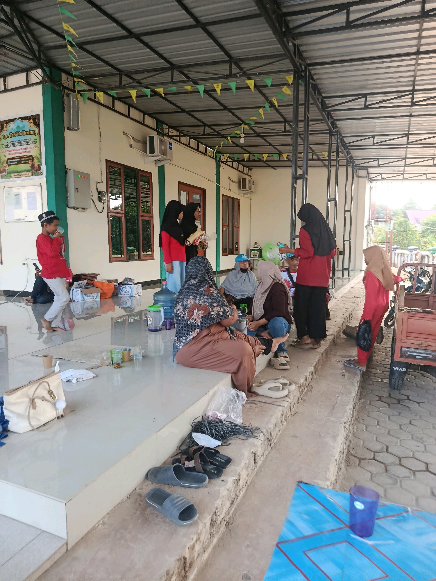 The image depicts an outdoor gathering at what appears to be a community center or school in Swarga Bara, Indonesia. The scene is set under a covered walkway with a metal roof supported by dark metal beams, decorated with strings of green and yellow triangular flags. The building behind has a light-colored exterior with green trim around the windows and the roofline.
Several people are present, mostly women and children, dressed in a mix of traditional and casual clothing. Some are wearing hijabs. One young boy in a red shirt and white pants is standing and appears to be drinking from a cup. Several individuals are sitting on the tiled floor or on raised concrete steps.
Scattered around the area are various objects, including several large blue water dispensers, plastic cups, some boxes, and bags. A light-colored handbag is placed on the floor near the raised steps, alongside some white fabric. Several pairs of sandals are also visible on the steps and the ground.
To the right of the frame, a small wooden cart with a red painted body is parked on a hexagonal paved surface. The overall atmosphere suggests a casual, possibly informal event or a waiting area. The lighting indicates it is daytime, with sunlight casting shadows.
