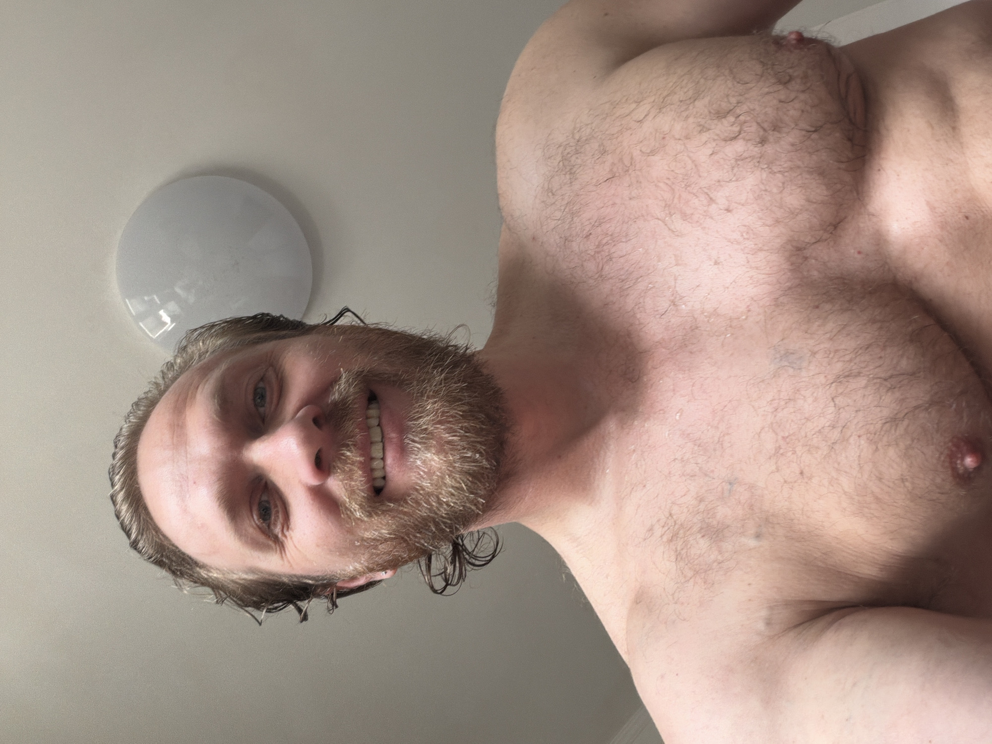 A selfie taken from a low angle depicts a shirtless man with fair skin, light brown hair, and a beard. His hair appears wet. The man is smiling, exposing his teeth. His chest is hairy. A round, white ceiling light fixture is visible in the upper left of the frame. The background is a plain, light-colored wall. There is no indication of this image being in Saint Petersburg, Russia.