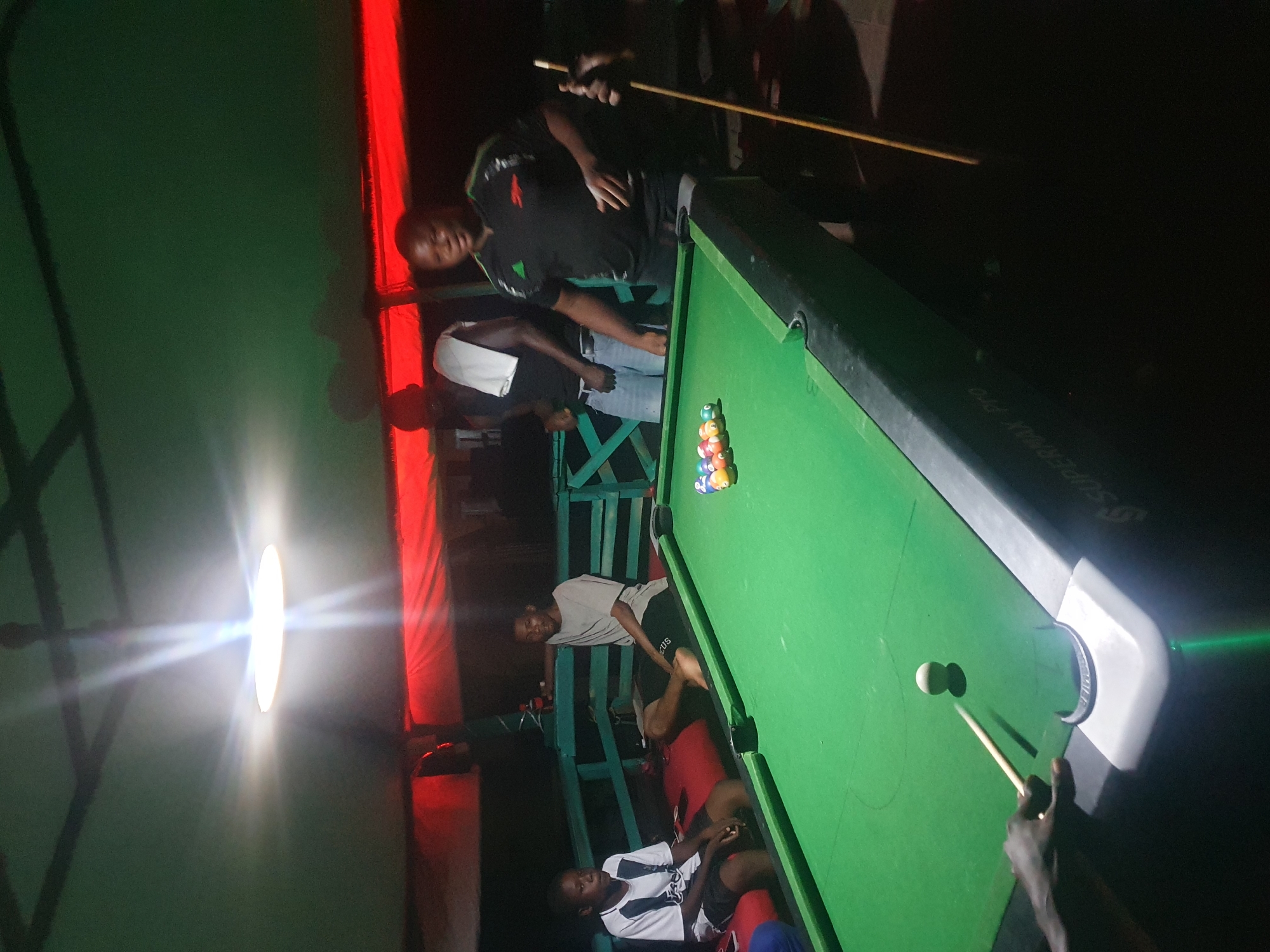 In Lokoja, Nigeria, a group of individuals are gathered around a pool table in what appears to be an outdoor or semi-outdoor entertainment area. The lighting is dim, with a bright overhead lamp casting strong shadows and a red glow emanating from above, possibly from decorative lights or a tent.
On the left, a young person wearing a black and white striped shirt and shorts is seated on a red bench, looking towards the pool table. Next to them, another young person in a grey t-shirt and dark shorts is also seated, with their legs extended.
In the center, a person wearing a black t-shirt with green accents is standing and holding a pool cue, appearing to be in the process of playing or about to play. To their left, another individual in a black t-shirt and light-colored shorts is standing, with a white towel draped over their shoulder.
The pool table itself is green, with a rack of colorful billiard balls at one end. One white cue ball is positioned on the table, with a pool cue aimed at it, suggesting an active game. The table has "SUPERMAX PRO" branding on its side. The surrounding environment includes wooden benches and railings, painted in a light blue-green color, and a dark, possibly canvas, ceiling. The overall atmosphere suggests a casual, recreational gathering.
