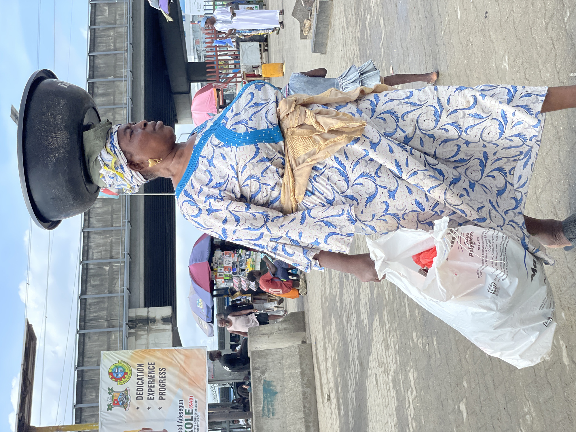 An older, dark-skinned African woman in Lekki, Nigeria is carrying a large black basin on her head. She is wearing a light-colored dress with blue patterns, a light-colored scarf, and gold earrings. She is also holding a white plastic bag in her hand. The background includes buildings, an advertisement reading "DEDICATION EXPERIENCE PROGRESS", vendors under umbrellas, and other people. The scene appears to be a bustling street.
