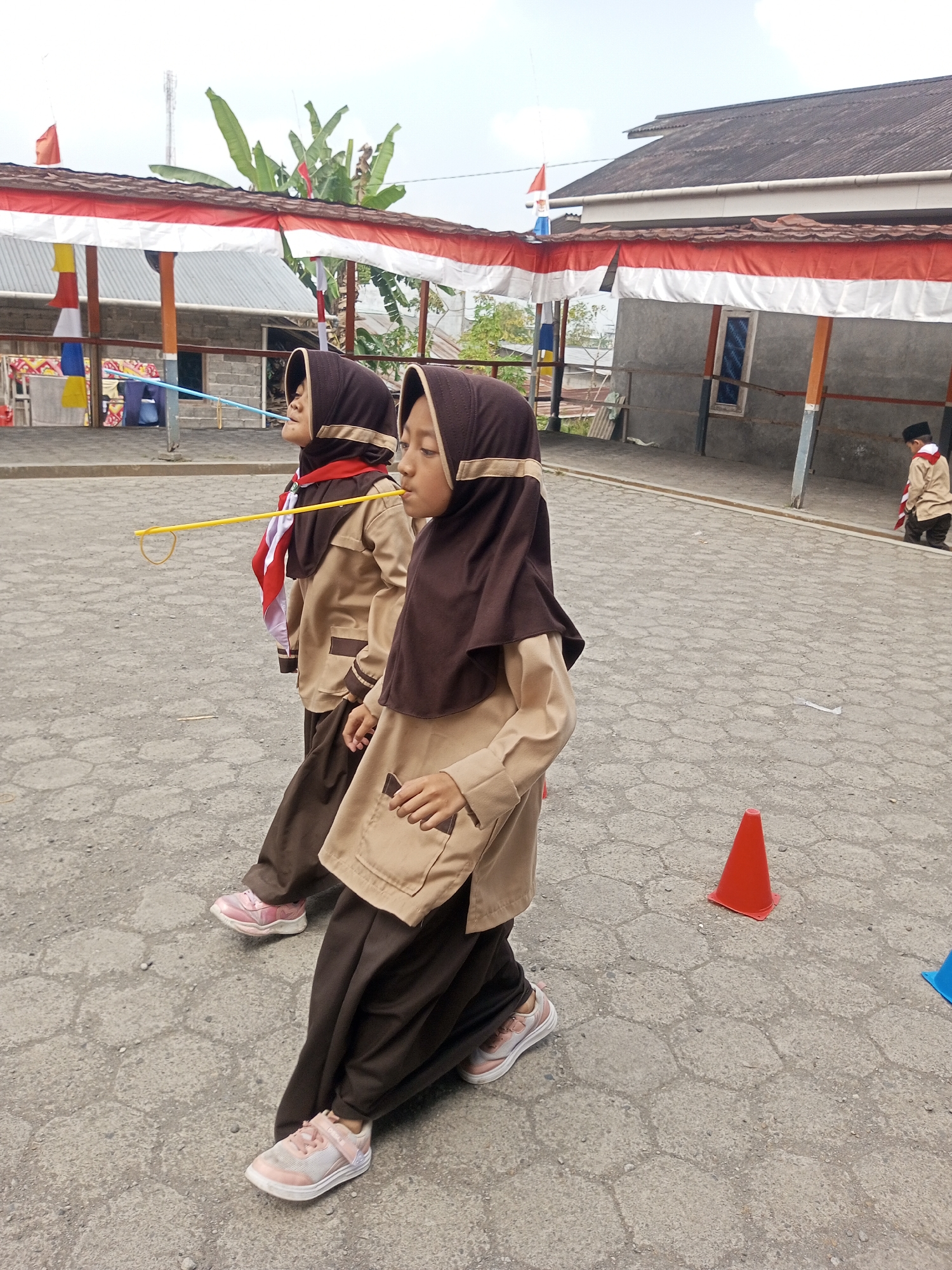 Two young girls, likely students in uniform, participate in an outdoor activity in Kalibeber, Indonesia. They are wearing tan uniforms with brown hijabs and scarves. The girl in the foreground is facing right, holding a yellow stick with a loop at the end in her mouth, and appears to be walking. The girl behind her is also facing right, with a similar yellow stick in her mouth, suggesting they are engaged in a relay or a game of some sort. The environment appears to be a paved area, possibly a schoolyard or community space, with a building in the background decorated with red and white banners. An orange traffic cone is visible on the ground to the right. The sky is overcast.