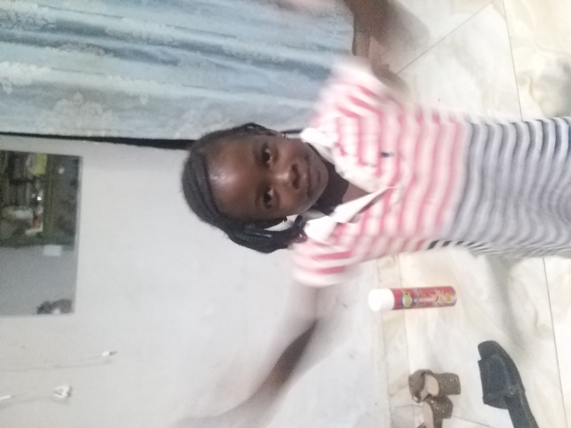 The image depicts a young Black girl with braided hair, standing indoors in front of a white wall and a blue curtain. She is wearing a horizontally striped shirt with shades of pink and gray. Her arms are blurred, suggesting movement. In the foreground, on the floor, are a pair of dark slide sandals, a tall cylindrical object that appears to be a can of spray, and a pair of sparkly heels. The floor is tiled with a marble-like pattern. The background shows a mirrored surface reflecting some indistinct objects, and the blue curtain is partially drawn. The setting appears to be a domestic interior, possibly a living room or bedroom, within the city of Kano, Nigeria.