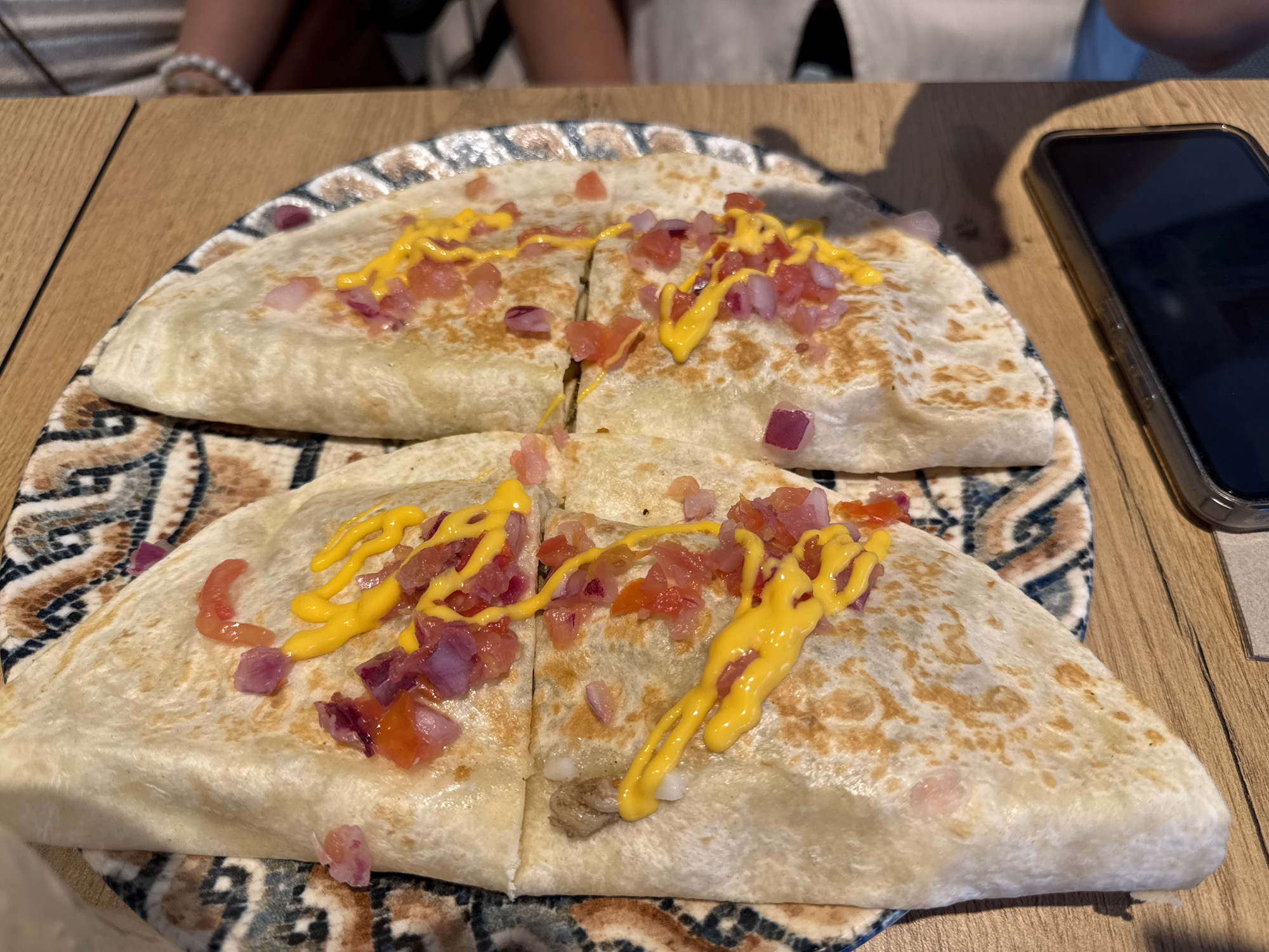 Two quesadillas, cut into quarters, are presented on a decorative plate. The quesadillas are topped with diced tomatoes, red onions, and a drizzle of yellow cheese sauce. A dark-colored smartphone is visible to the right of the plate. The scene appears to be set on a wooden table, possibly in a restaurant. The background shows the blurry figures of at least two people and a hint of what might be a menu or a white napkin. The lighting is natural and casts shadows on the table and plate. The location is specified as Aldaia, Spain.