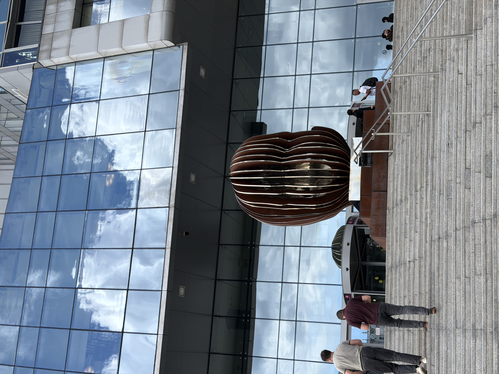 The photograph shows a modern architectural scene in Moscow, Russia. A spherical sculpture composed of brown, horizontally-oriented slats stands near the entrance of a building with a mirrored facade, reflecting the sky. People are present: two individuals are walking on a gray, tiled walkway leading towards the entrance, and a small group is positioned at the top of a staircase. The lighting suggests daylight.