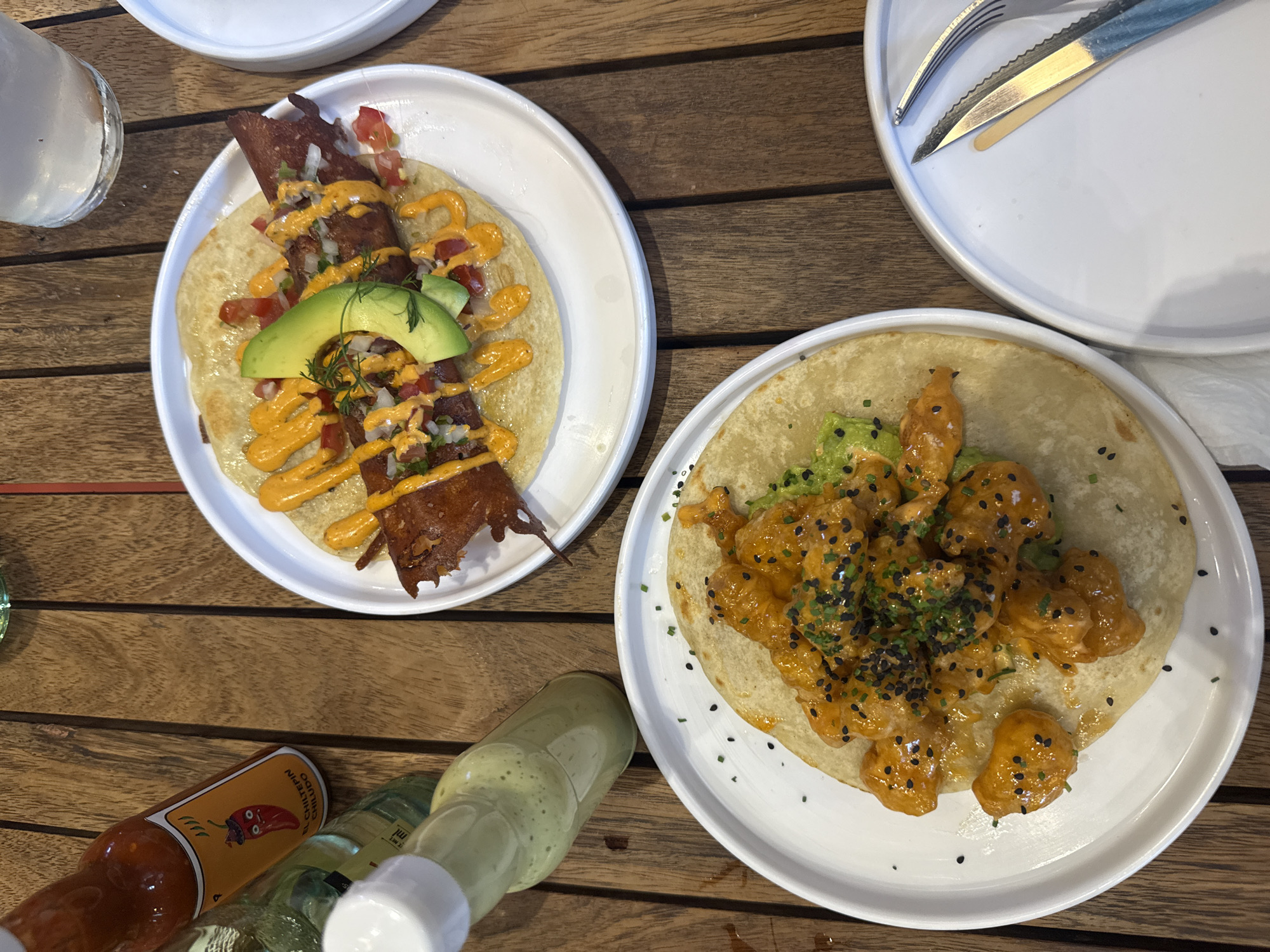 The image depicts a wooden table setting, featuring two white plates each holding a taco. The taco on the left plate is topped with a yellow sauce, avocado slices, diced vegetables, and large, dark brown, rectangular slices of what appears to be meat or a meat substitute. The taco on the right plate features a similar yellow sauce, avocado, and black sesame seeds.
A glass of liquid is partially visible on the left side of the frame, near the plate with the meat-like topping. To the right, on top of another white plate, there are a knife and fork set. At the bottom, several bottles of hot sauce are present.
Given the type of tacos and presentation, it is likely the location is Mexico City, Mexico.