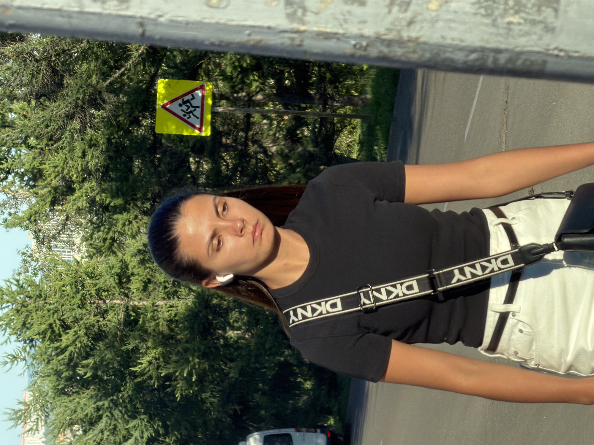An outdoor medium shot captures a young woman walking, her body angled slightly to the left, her face turned towards the viewer. She wears a black t-shirt, white shorts, and a black crossbody bag with a white DKNY strap. White wireless earbuds are visible in her ears. Behind her, a dense line of green trees dominates the background. A yellow triangular traffic sign with a red border, depicting figures running, is positioned in the middle ground, to the right of the woman. The scene appears to be a street in Krasnoyarsk, Russia, indicated by the presence of urban vegetation and residential buildings visible in the distance through the trees. The lighting suggests it is daytime, with sunlight casting shadows.